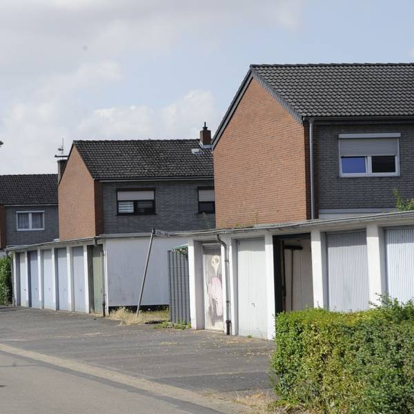 Zu sehen ist eine Straße mit Garagen und parkenden Autos davor, dahinter sind Häuser zu sehen.