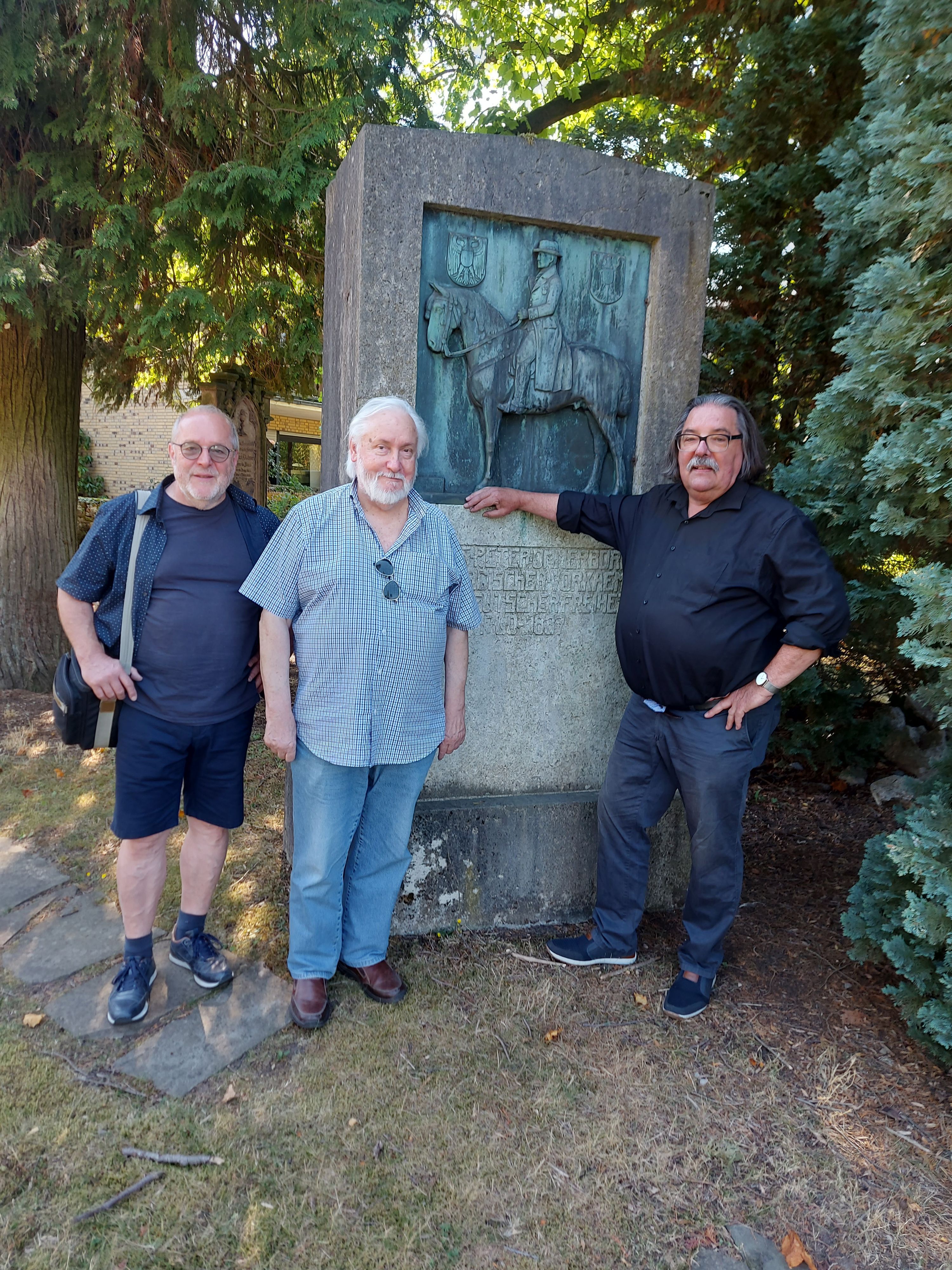 Drei Männer stehen vor einem Denkmal mit Bronzeplakette. Darauf ein Mann zu Pferd.