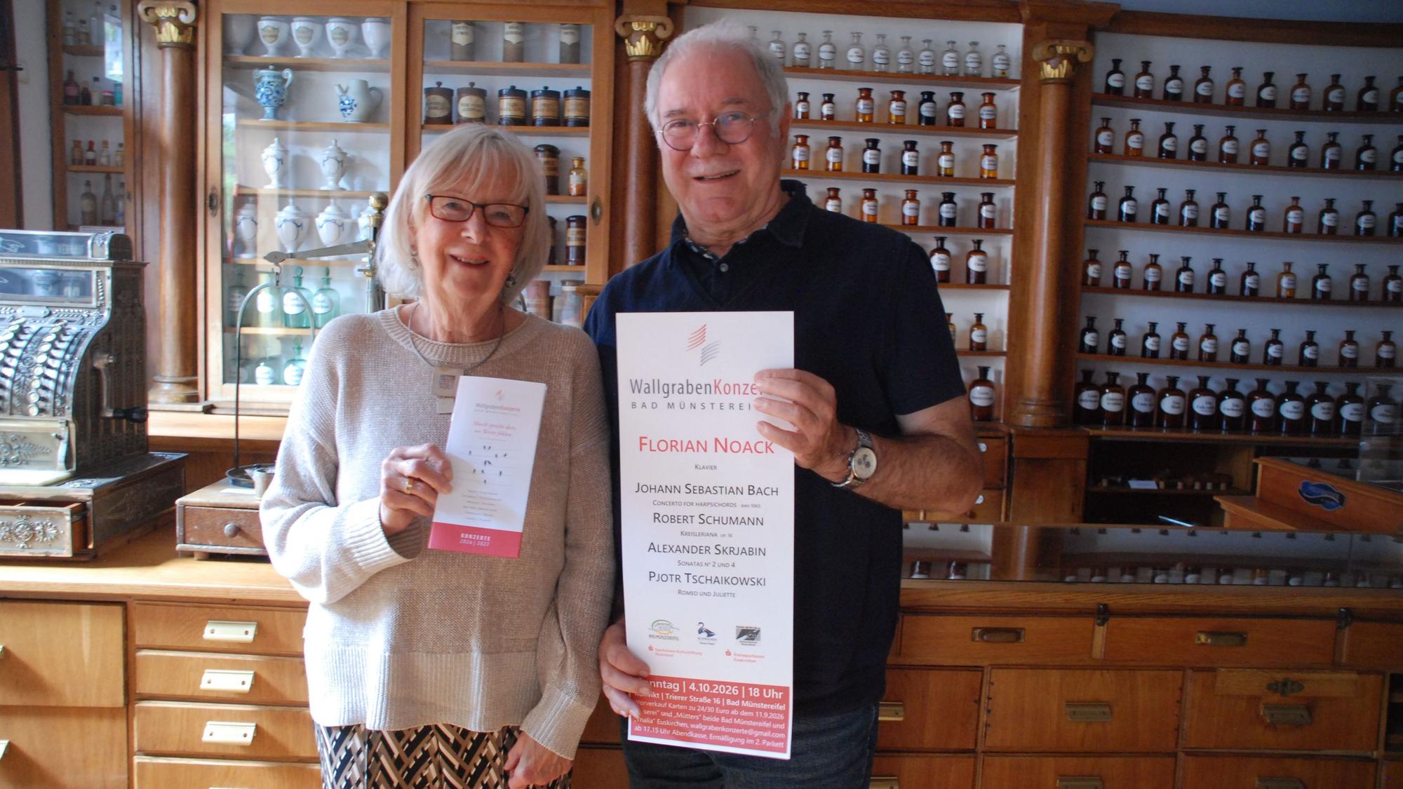 Eine Frau und ein Mann stehen vor einer Wand mit Apothekenfläschen und halten ein Plakat und einen Flyer in der Hand.