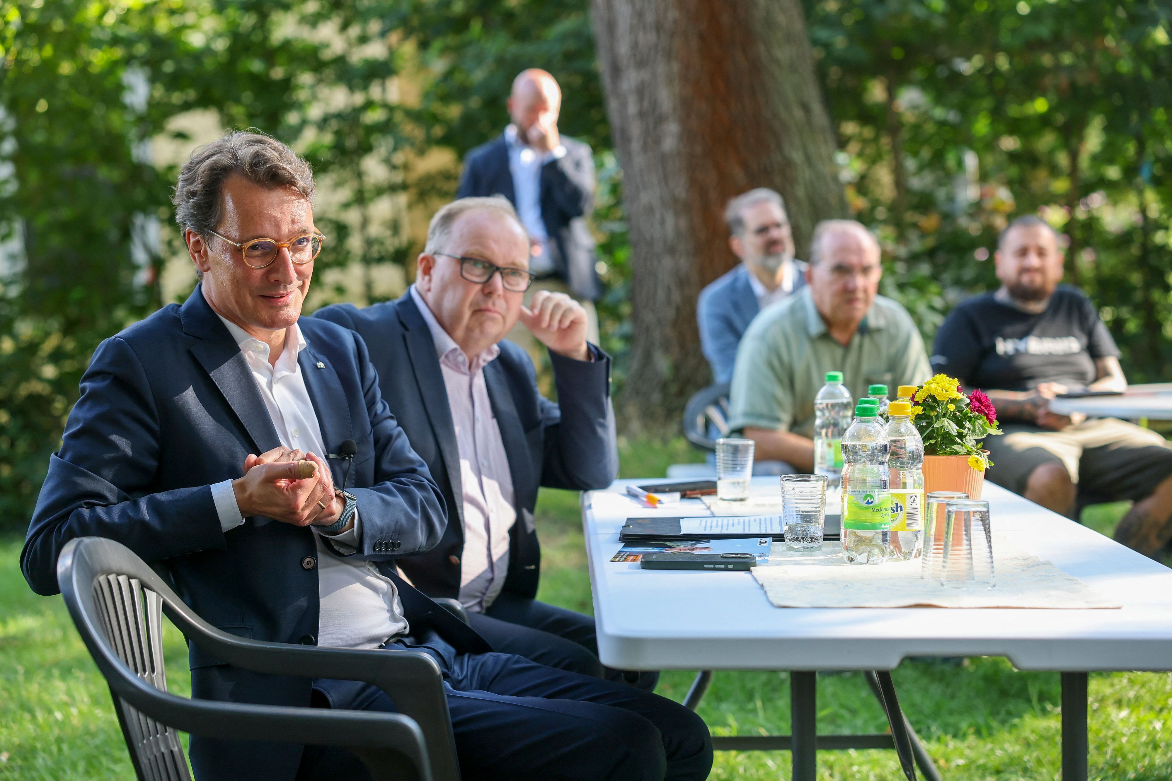 Hendrik Wüst (l, CDU), Ministerpräsident von Nordrhein-Westfalen, und Michael Scheffler (CDU), Landtagsabgeordneter Sachsen-Anhalt, sitzen im Garten eines Dorfgemeinschaftshauses und sprechen mit Bürgern.