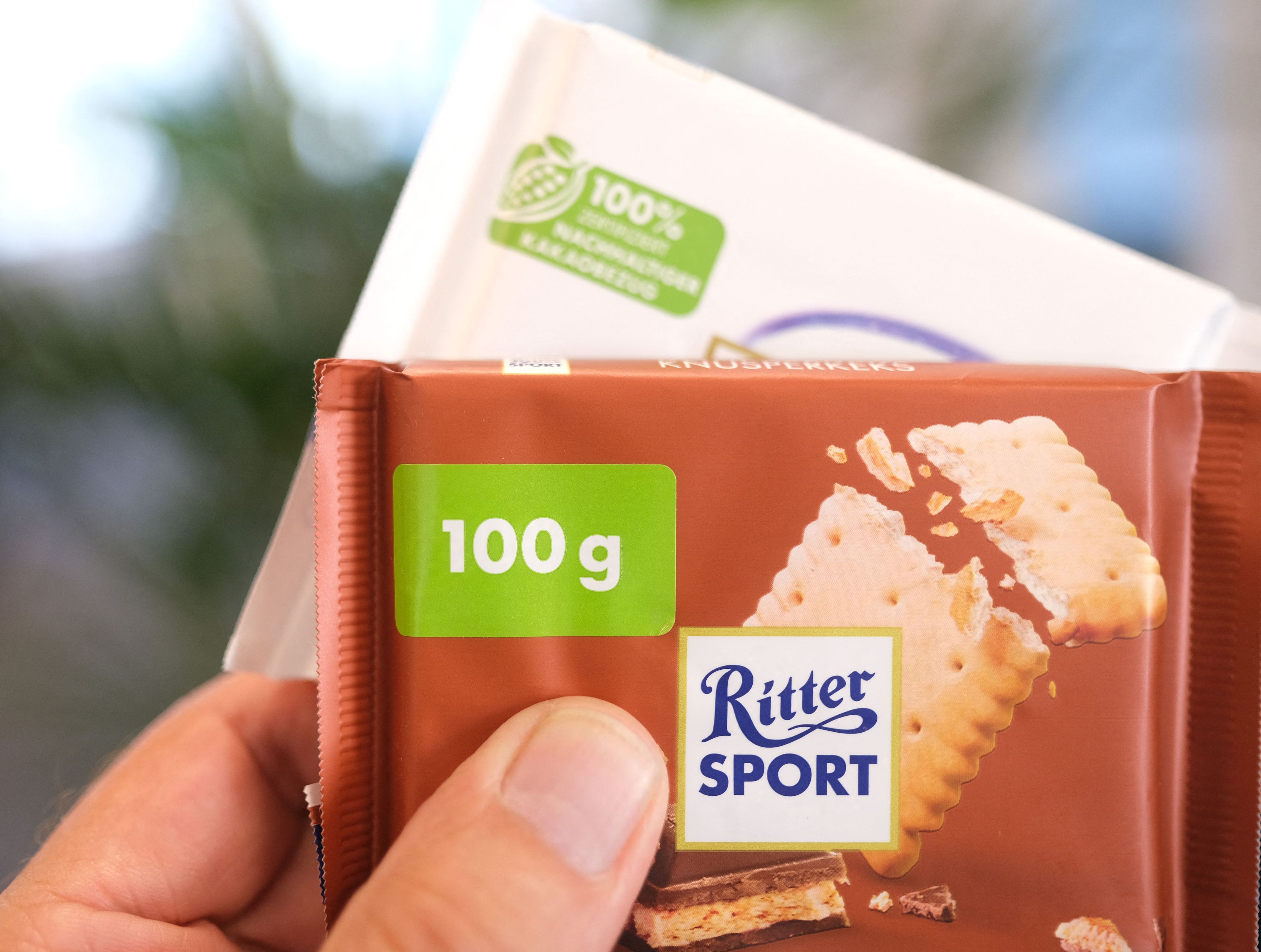 Eine Hand hält zwei Tafeln Schokolade von Ritter Sport. Auf der vorderen Tafel ist auf der Verpackung ein Hinweis mit einem Sticker überklebt.