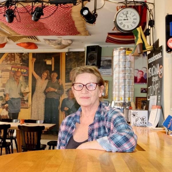 Eine Frau mit kurzem Haar, Brille und Karo-Hemd sitzt an der Bar in einer geräumigen Kneipe.