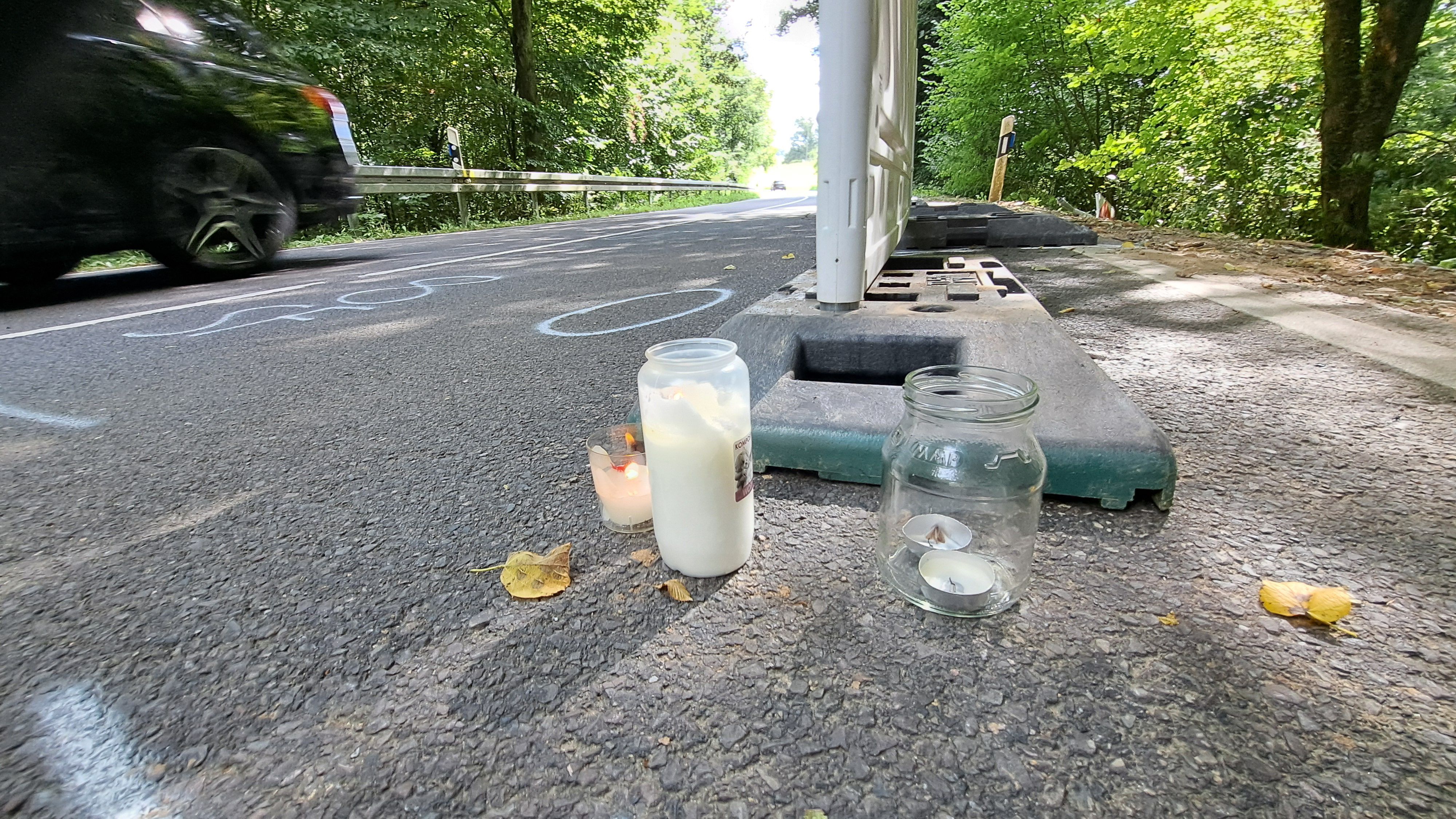 Eine Straße mit Grabkerzen rechts vor einer Bake und links einem vorbeifahrenden Auto.
