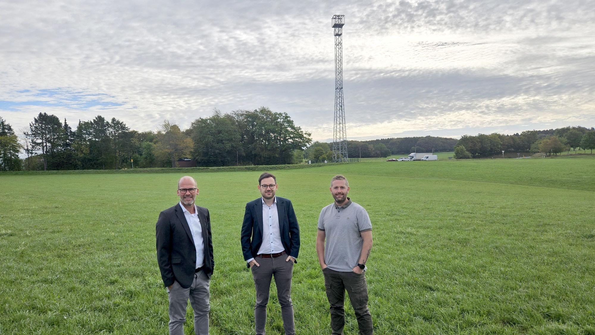 Drei Männer stehen nebeneinander auf einer Wiese. Im Hintergrund ist der Funkmast zu sehen.