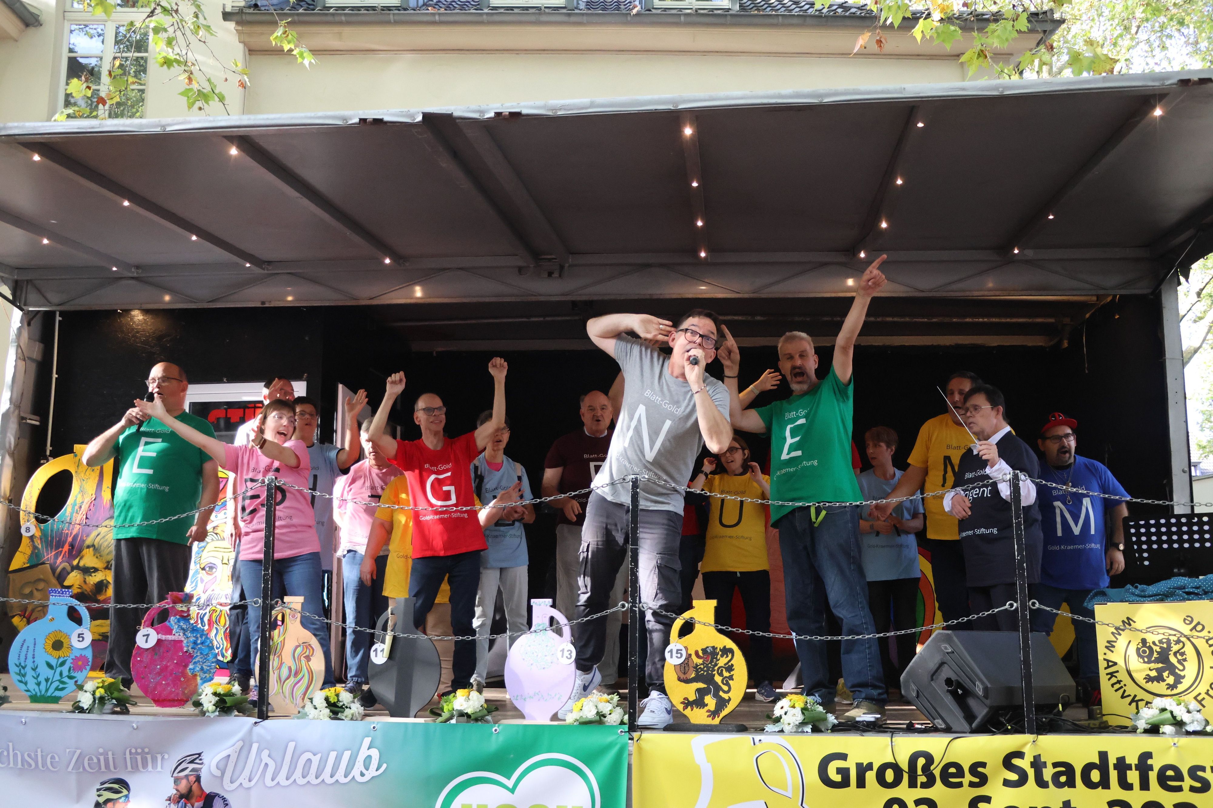 Auf dem Bild sind Chormitglieder in bunten T-Shirts auf der Bühne zu sehen.