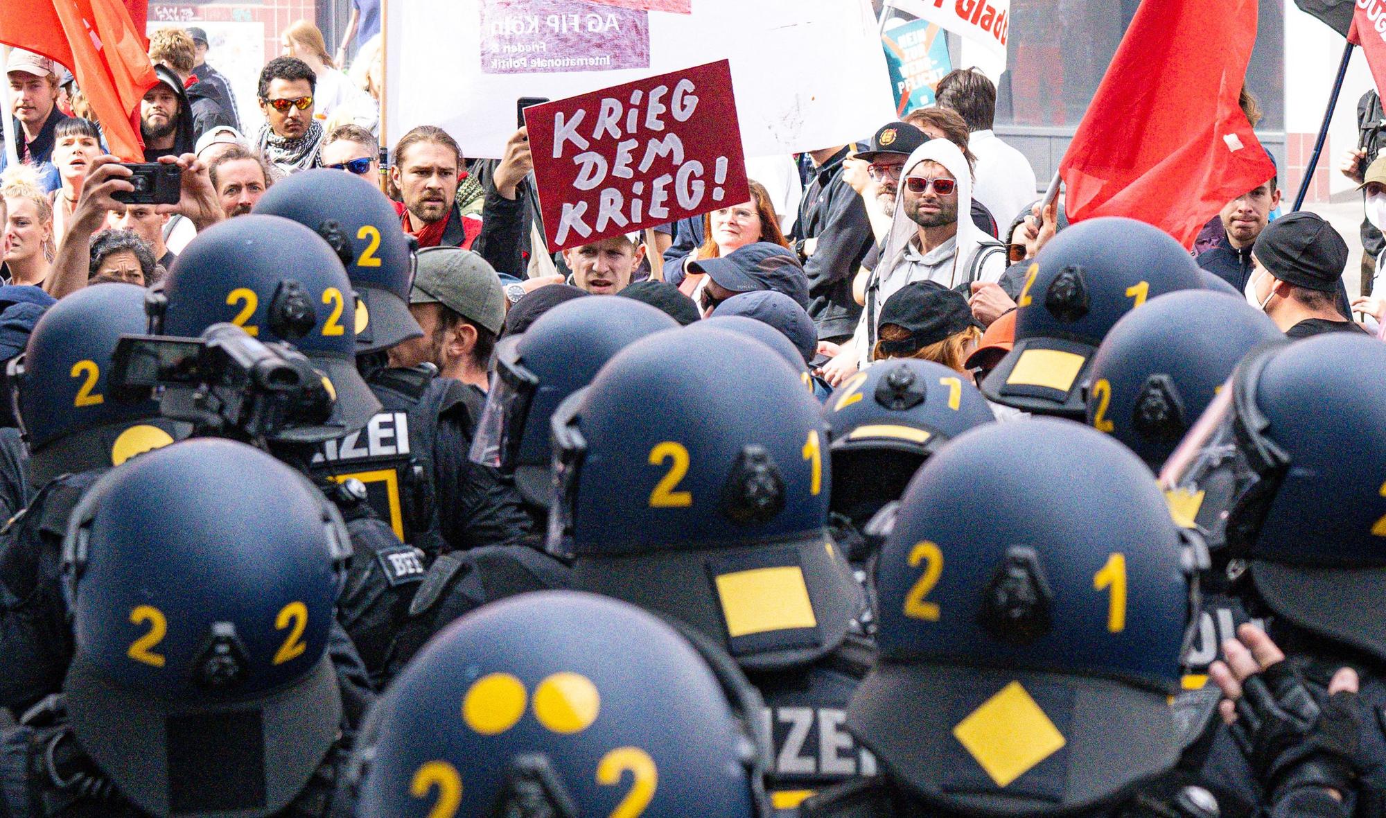 Bei der Abschlussdemonstration des Protestcamps „Rheinmetall entwaffnen“ hält ein Teilnehmer der Demo ein Schild mit der Aufschrift „Krieg dem Krieg“. Im Vordergrund sind Polizeikräfte zu sehen.