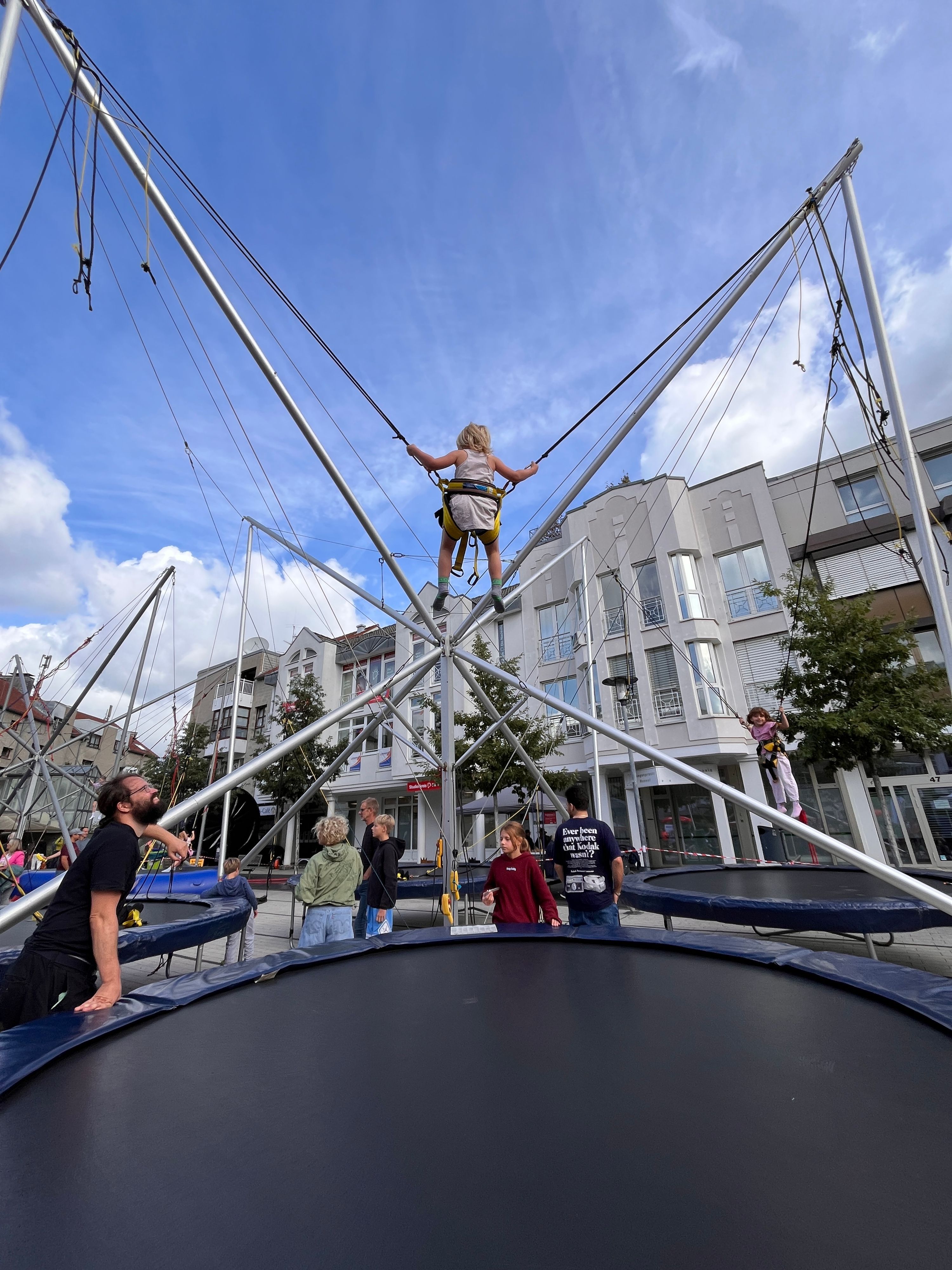 Ein kleines Mädchen schwingt in einem Tragesitz über einem Trampolin auf und ab.