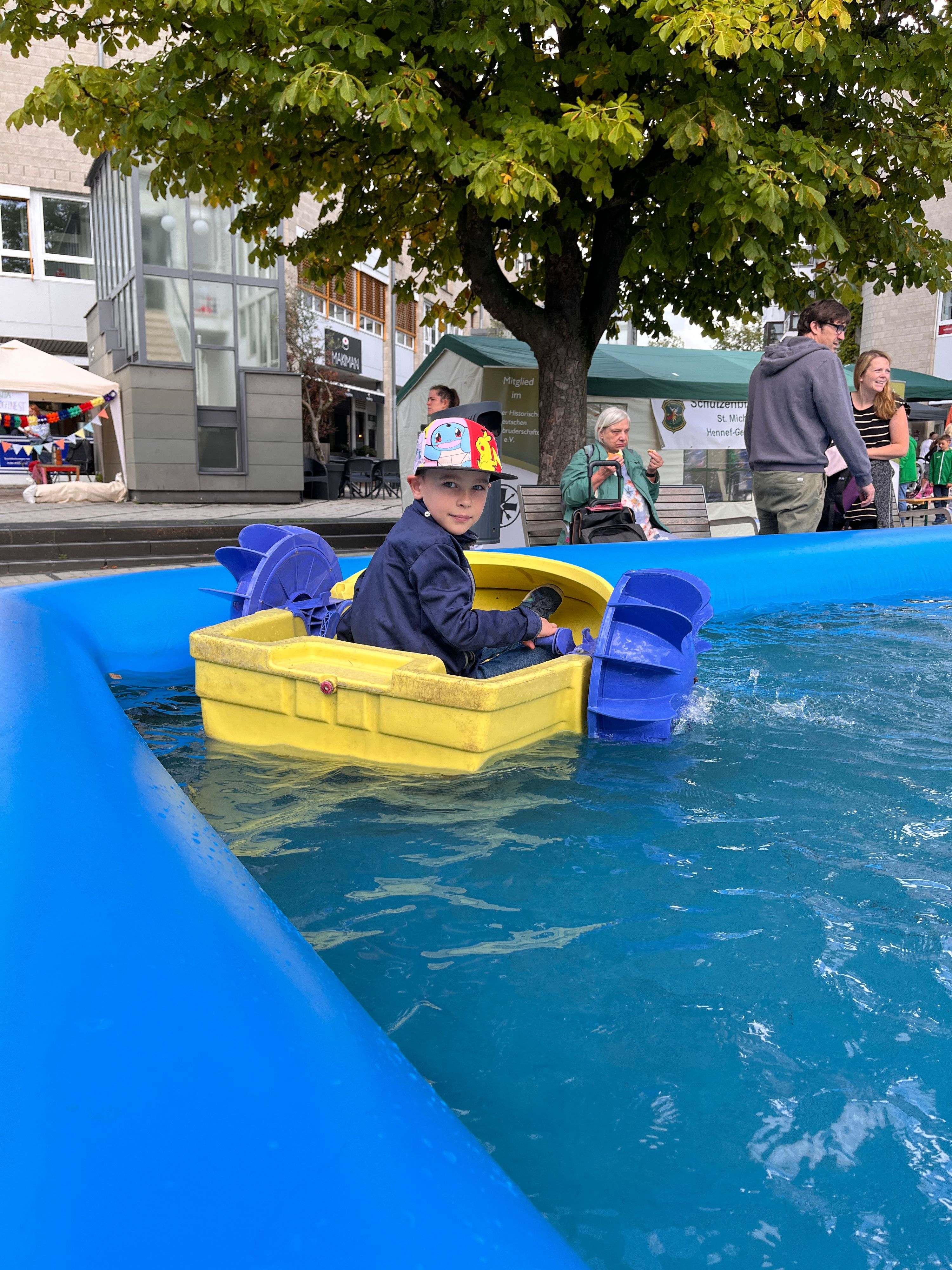 Ein Junge mit Baseballkappe sitzt in einem kleinen Boot mit Schaufelrädern, das in einem Wasserbecken schwimmt.