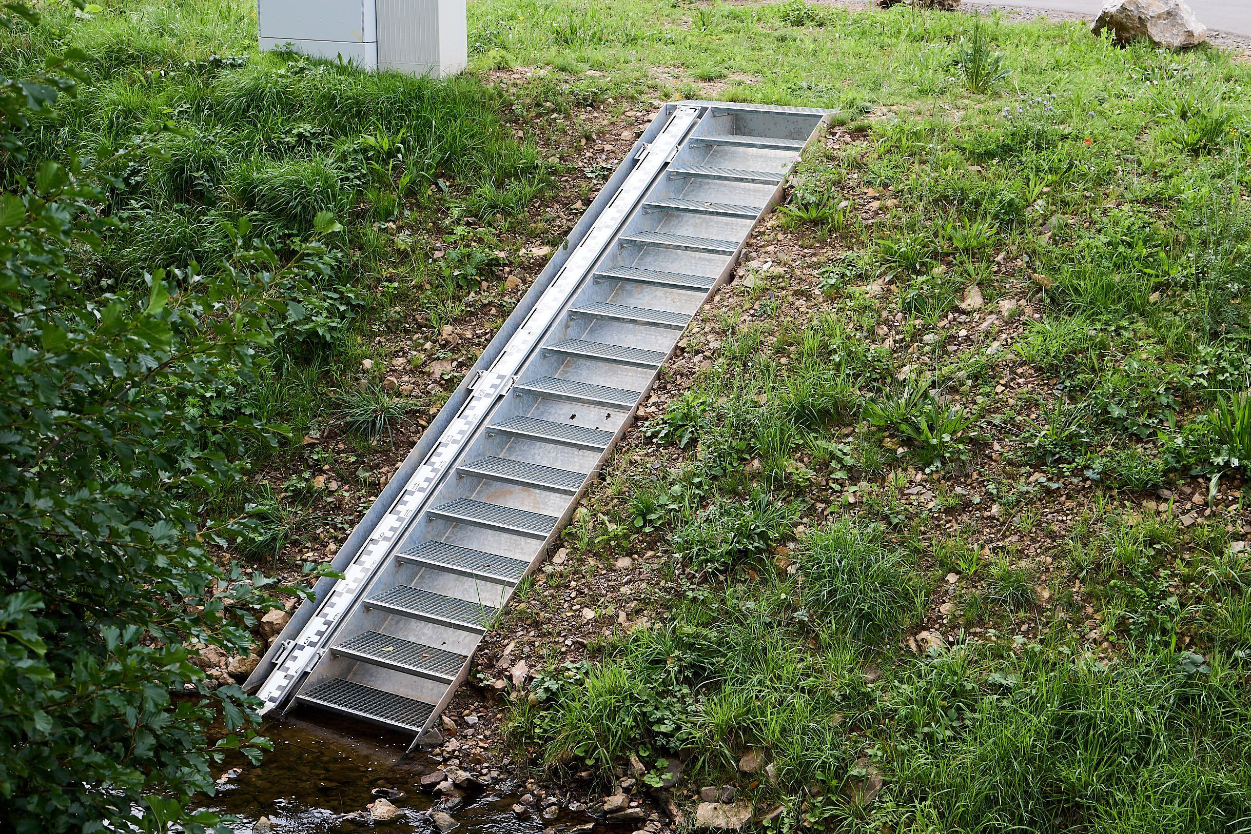 Der Pegel ist in die Bachböschung eingebaut worden, er wird von einer Metalltreppe flankiert.