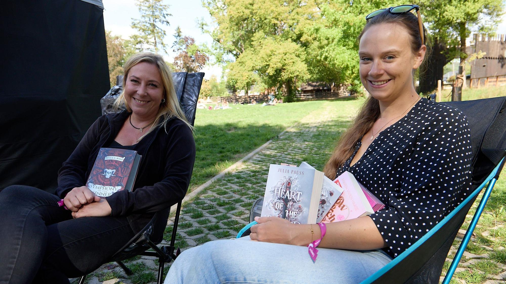 Zwei Frauen sitzen in Klappstühlen auf einer Wiese. Sie halten Bücher in den Händen.