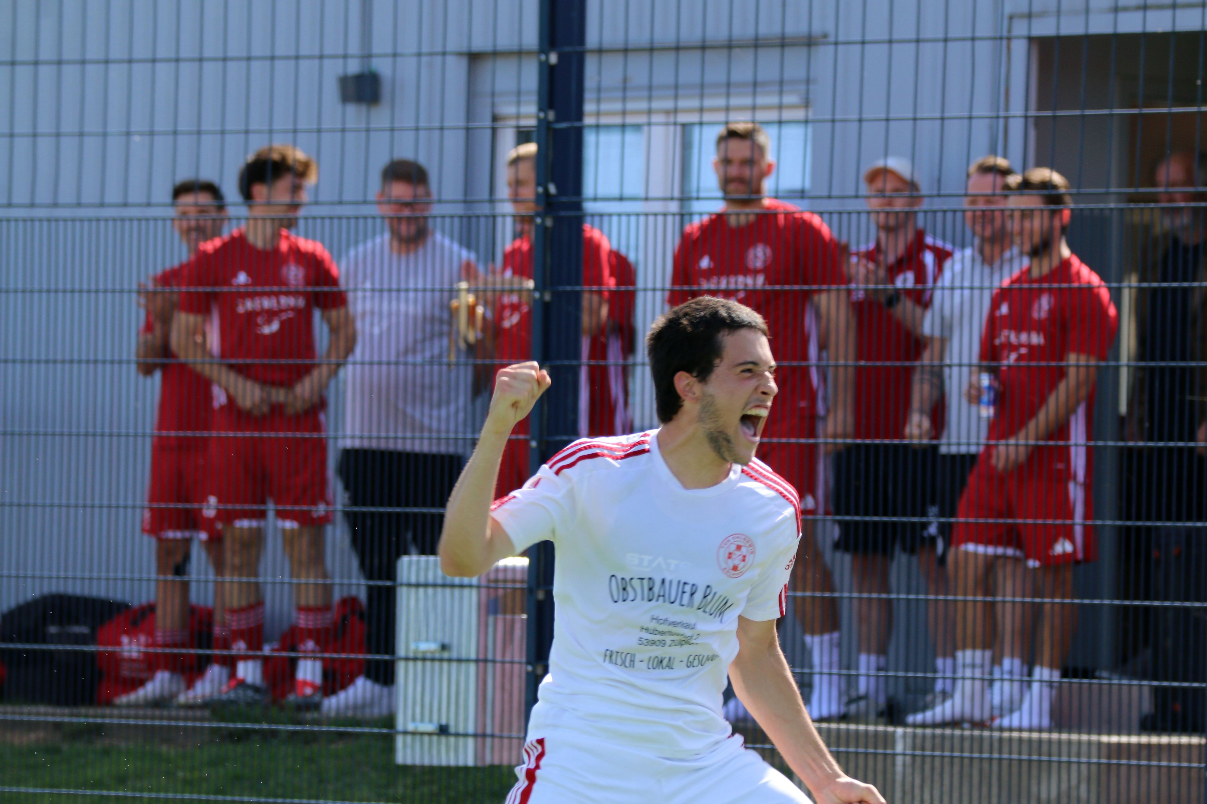 Ein Zülpicher Spieler jubelt und hält die Faust in die Höhe. Im Hintergrund stehen Spieler und Betreuer der ersten Mannschaft und schauen zu.