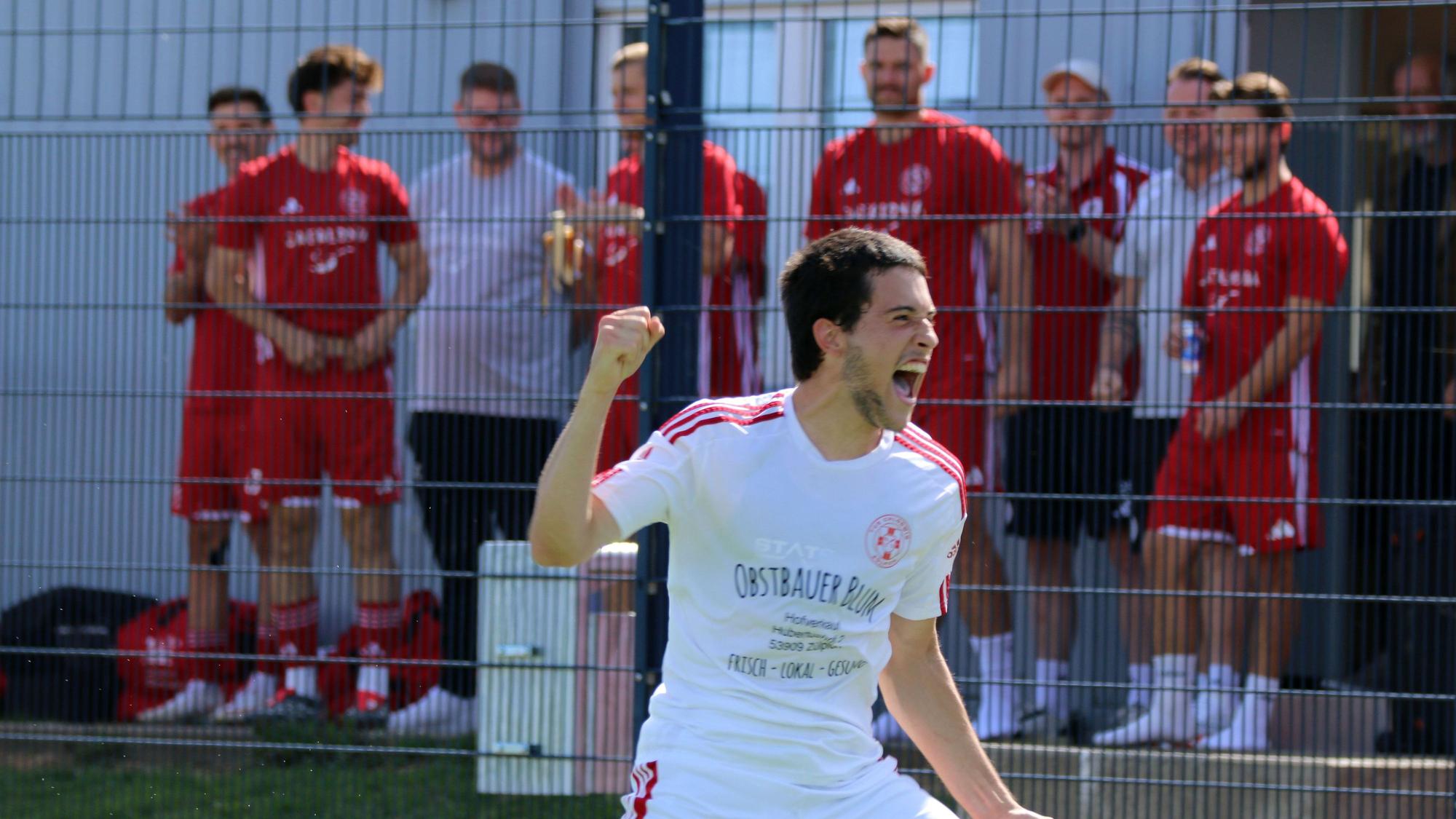 Ein Zülpicher Spieler jubelt und hält die Faust in die Höhe. Im Hintergrund stehen Spieler und Betreuer der ersten Mannschaft und schauen zu.