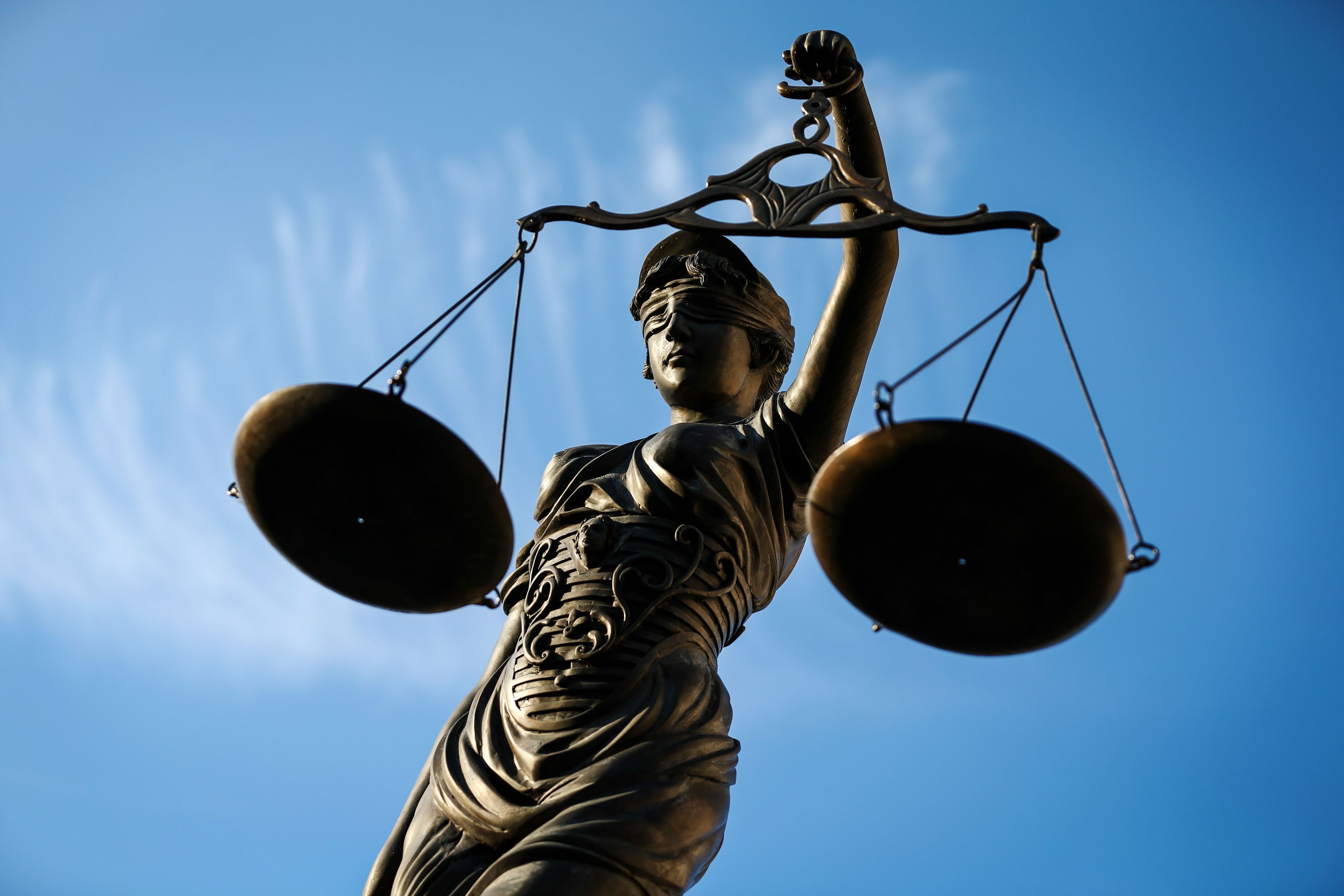 Eine Justitia aus Bronze mit verbundenen Augen hält zwei Waagschalen gegen einen blauen Himmel.
