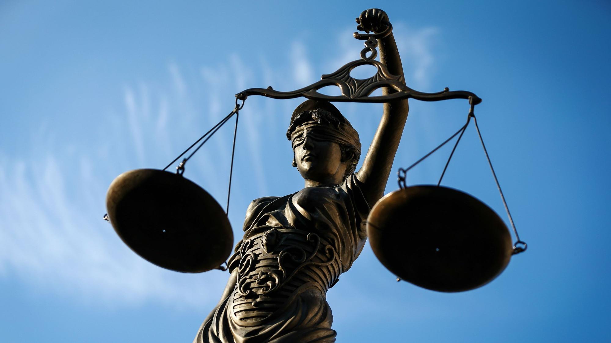 Eine Justitia aus Bronze mit verbundenen Augen hält zwei Waagschalen gegen einen blauen Himmel.