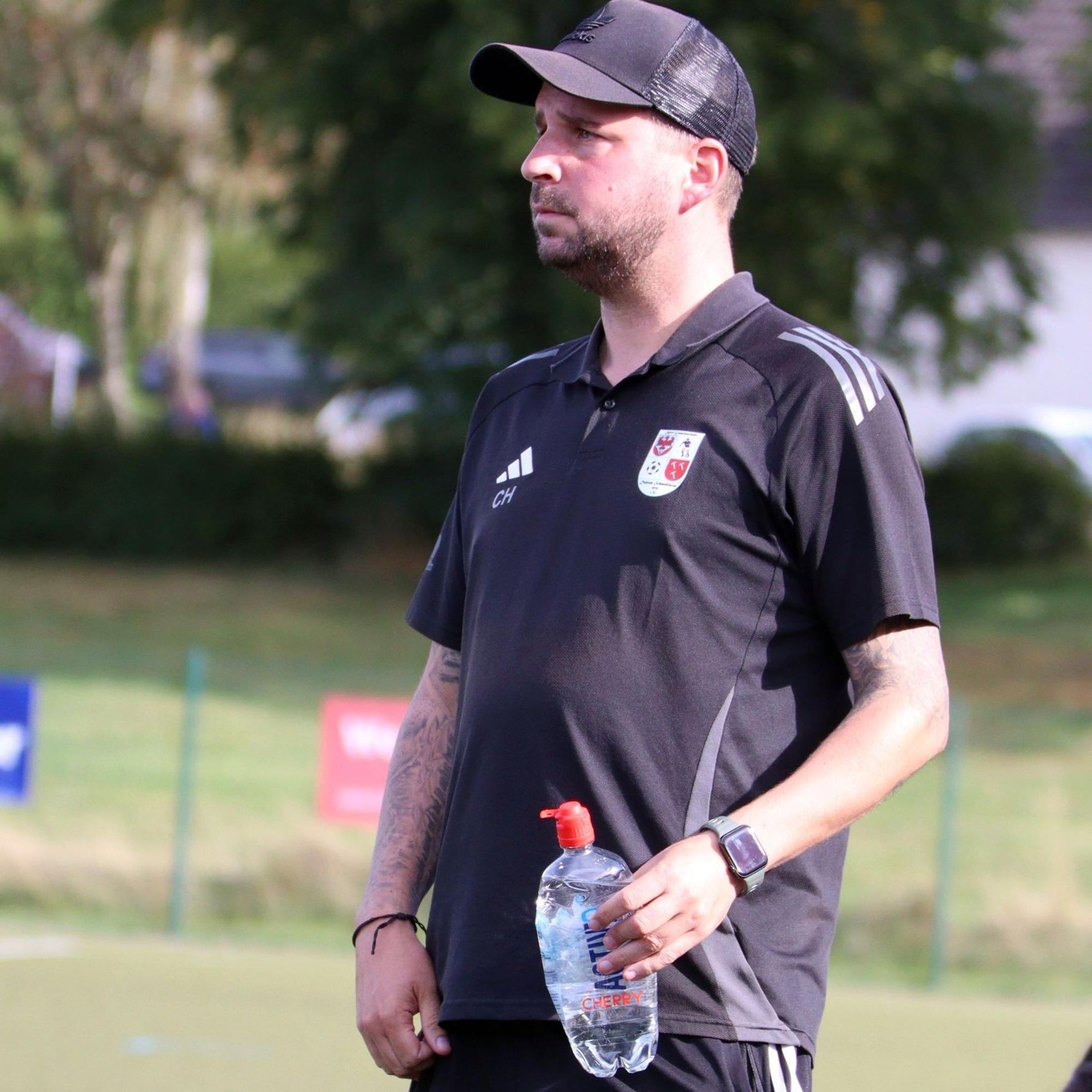 Dahlems Trainer Christian Hammes hält in einer Hand eine Wasserflasche und schaut dem Spiel zu.