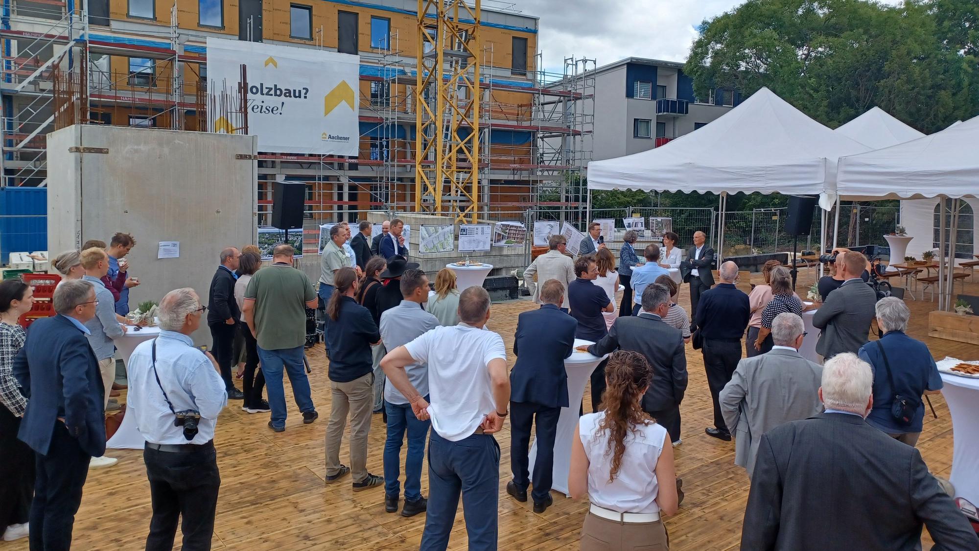 Viele Menschen stehen auf einer Baustelle. Im Hintergrund sind ein eingerüsteter Rohbau und ein Kran zu sehen.
