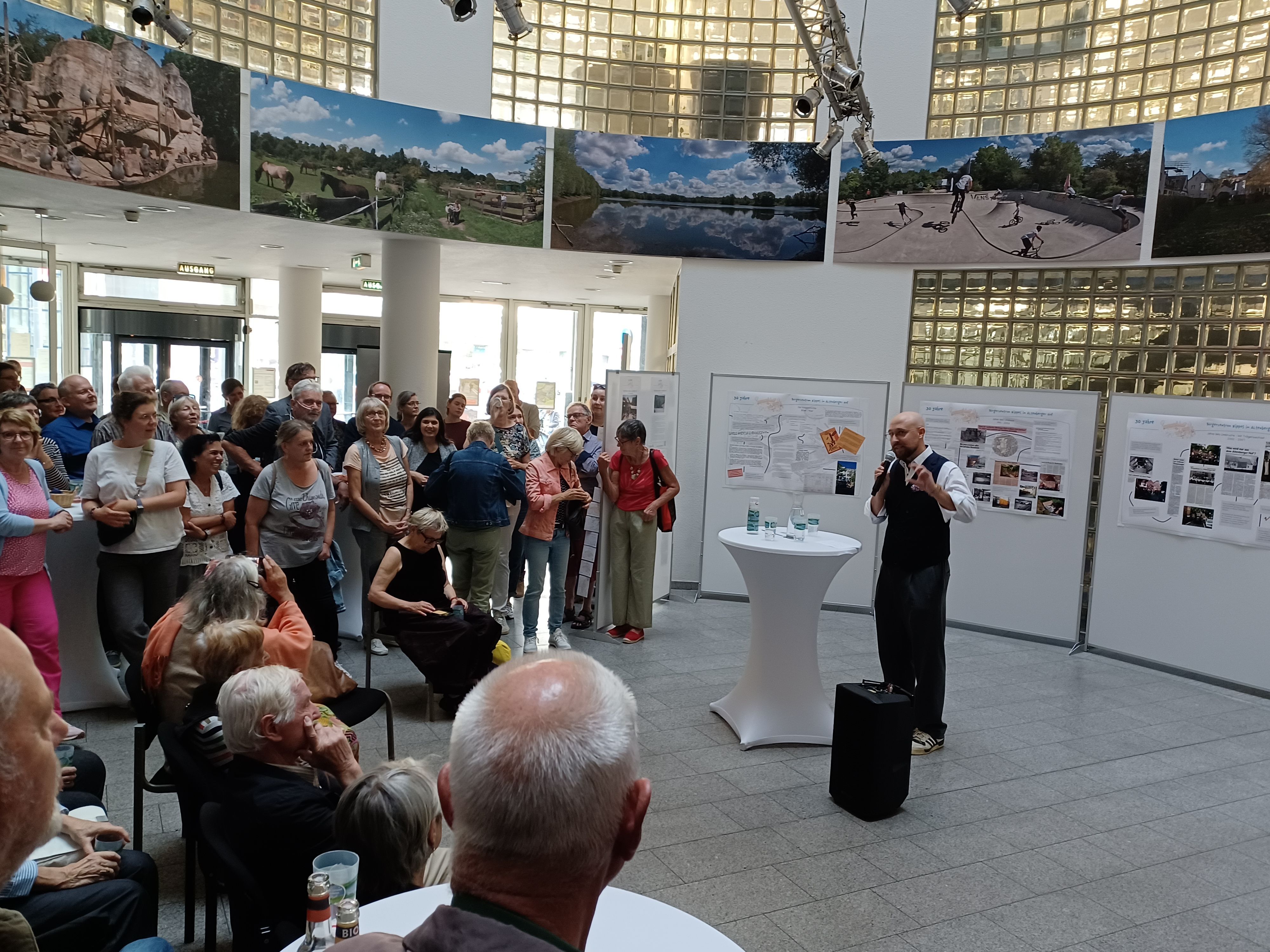 An einem Stehtisch vor Ausstellungswänden steht ein Mann mit Glatze. Er spricht zu Menschen, die im Halbkreis vor ihm stehen oder sitzen.