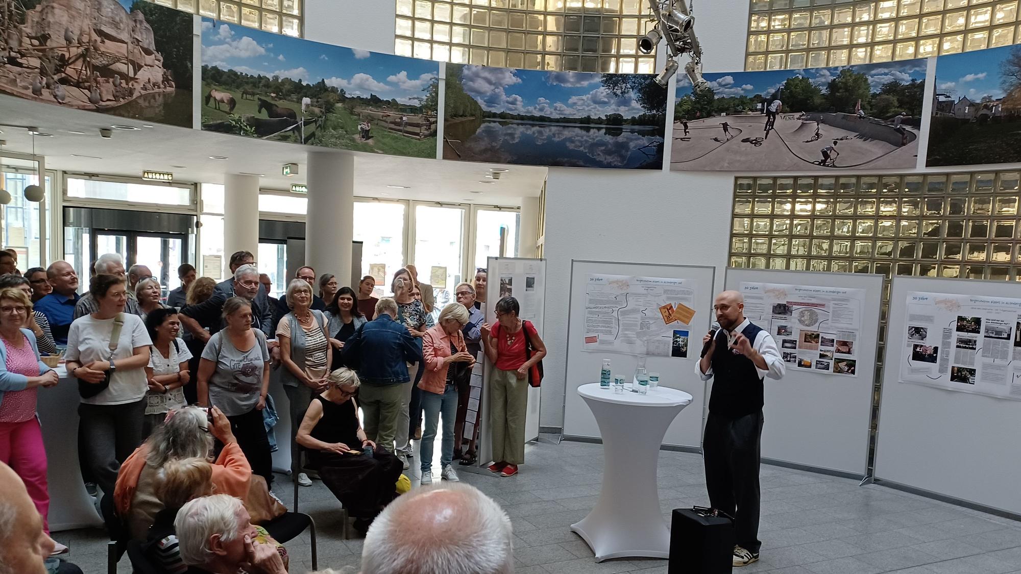 An einem Stehtisch vor Ausstellungswänden steht ein Mann mit Glatze. Er spricht zu Menschen, die im Halbkreis vor ihm stehen oder sitzen.