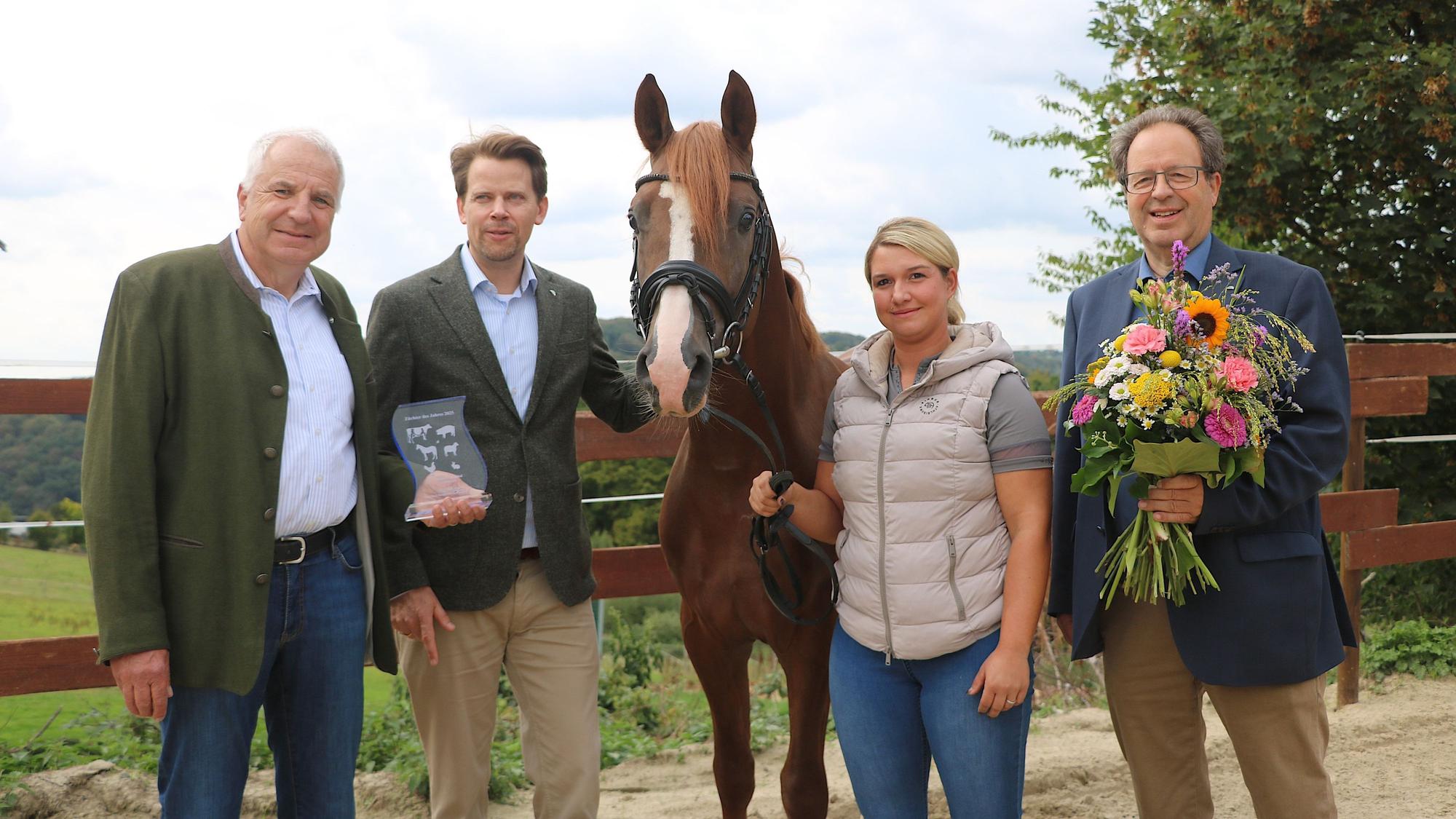 Eine blonde Frau hält ein braunes Pferd am Zügel, daneben stehen drei Männer, einer mit Blumenstrauß.