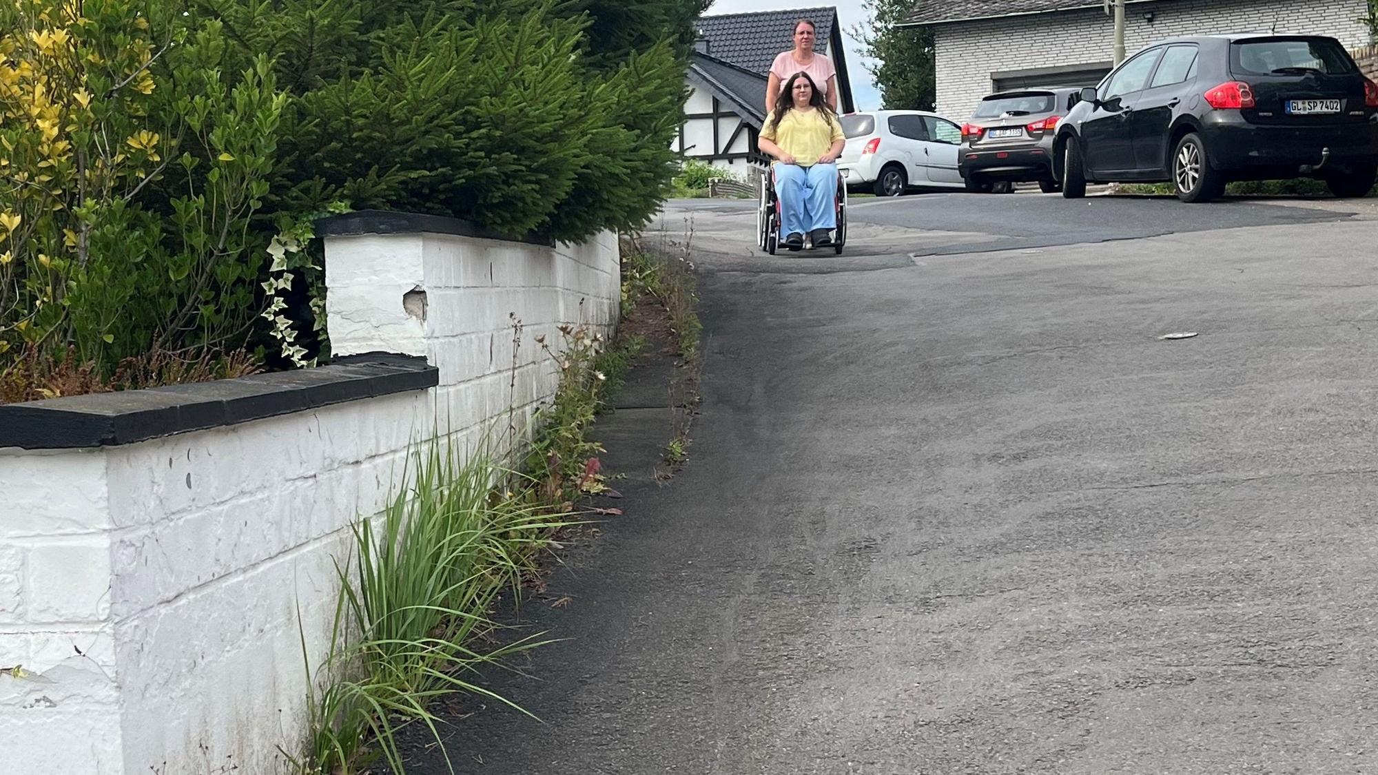 Die junge Frau wird im Rollstuhl von ihrer Mutter geschoben.