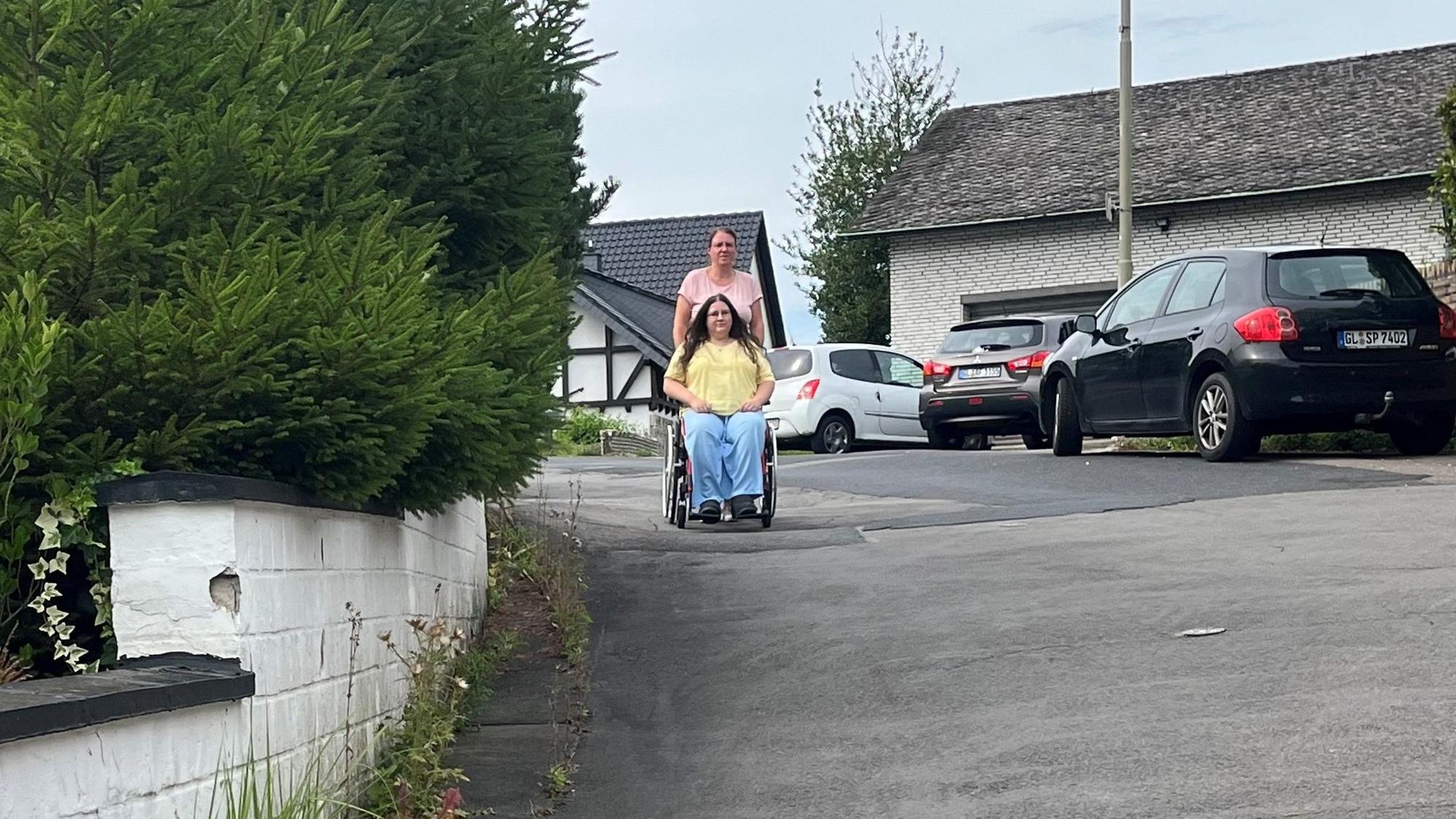 Die junge Frau wird im Rollstuhl von ihrer Mutter geschoben.