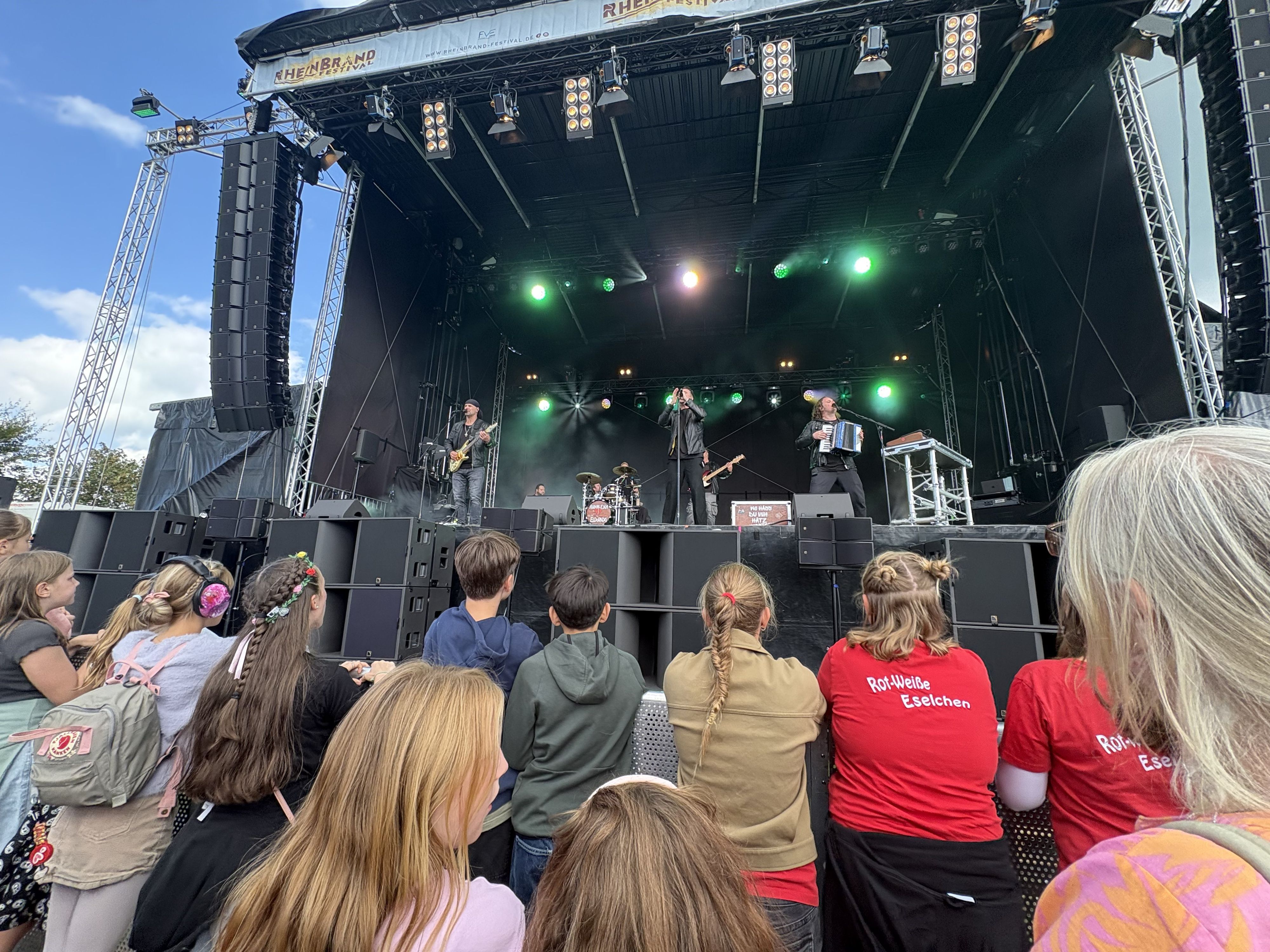 Jugendliche stehen vor einer großen Bühne. Dort spielt eine Band.