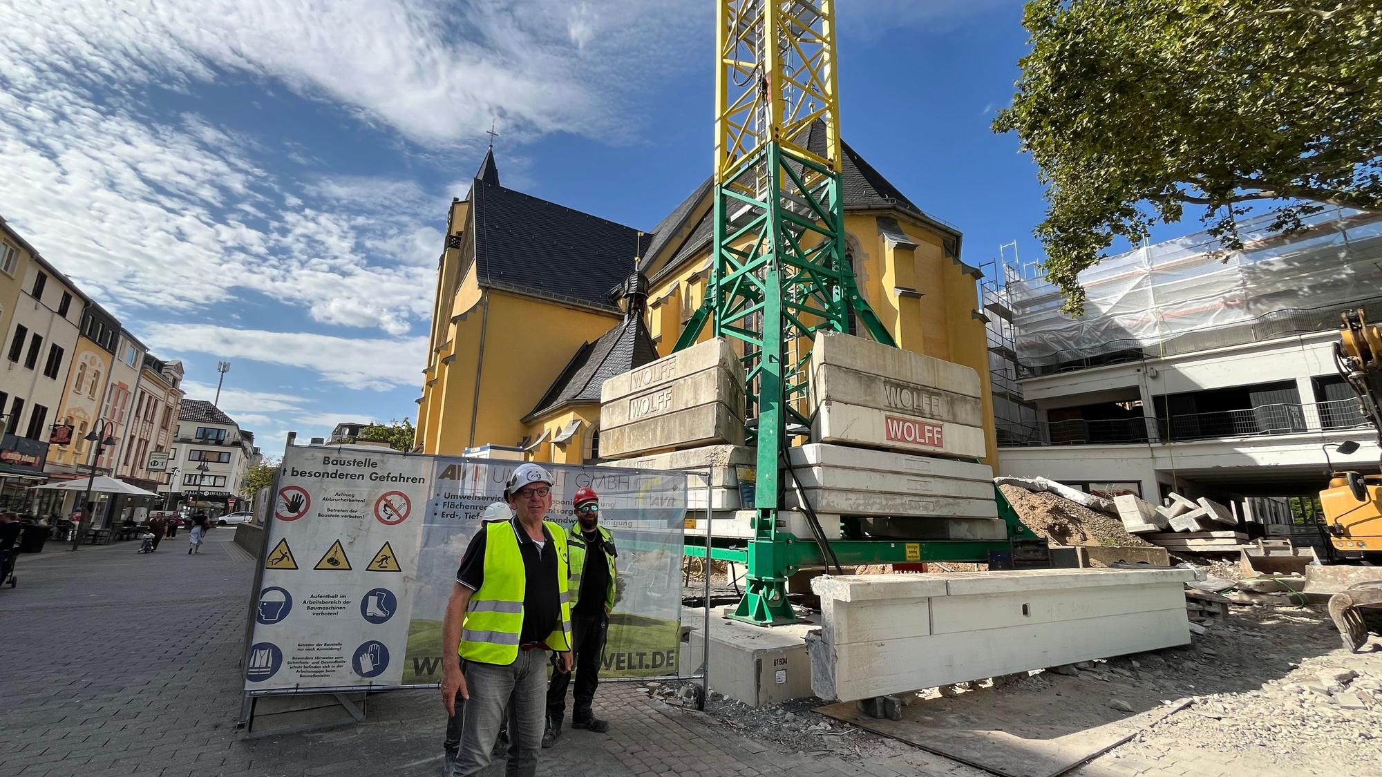 Ein Kran und schwere Betonteile stehen vor einer Kirche in der Fußgängerzone von Porz.