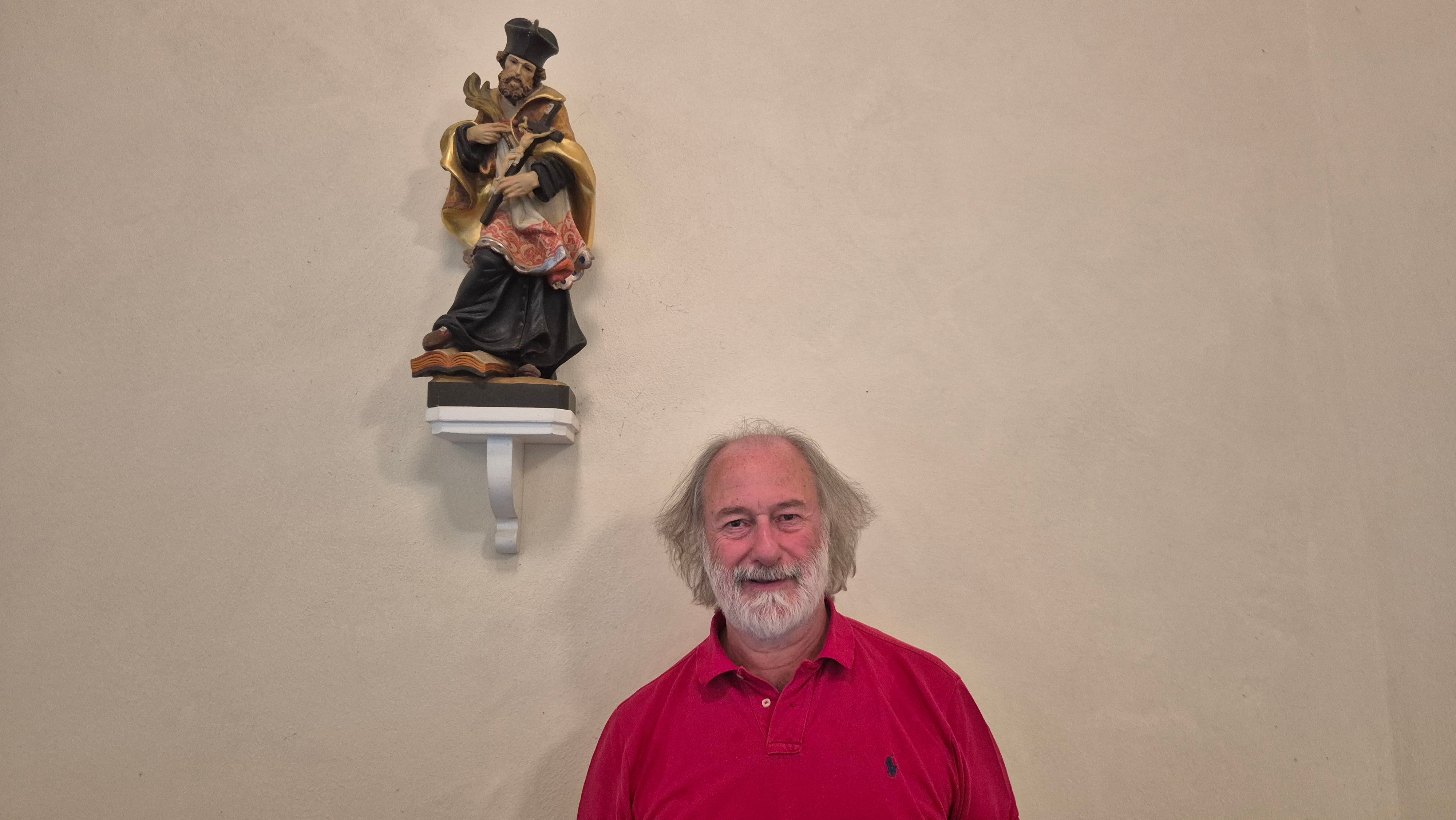 Ein Mann im roten Polohemd steht vor einer Kirchenwand, an der eine sakrale Statue auf einem Sockel steht.