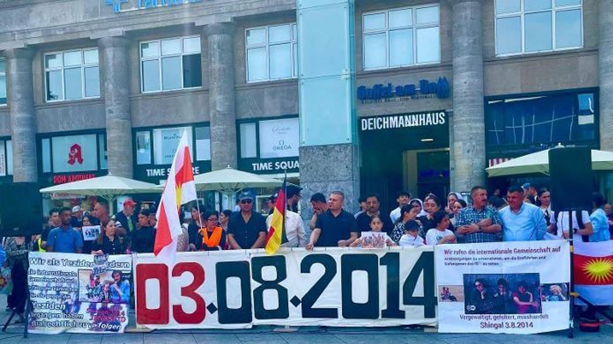Menschen stehen hinter einem Banner, das an das Massaker an den Jesiden am 3.8. 2014 erinnert.