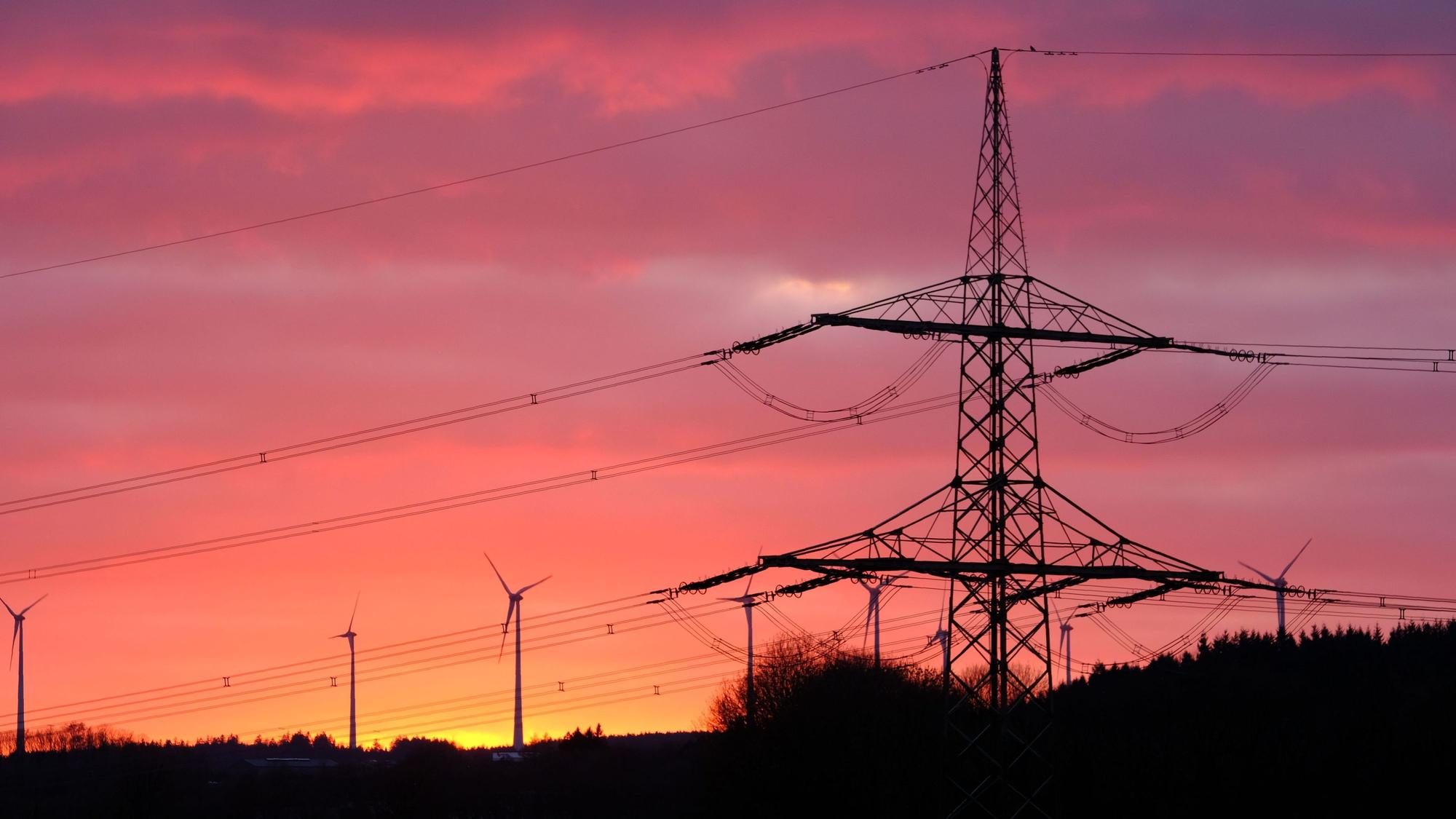 Der Mast ener 110-kV-Höchstspannungsleitung und mehrere Windkraftanlagen im Abendrot bei Dahlem-Schmidtheim.