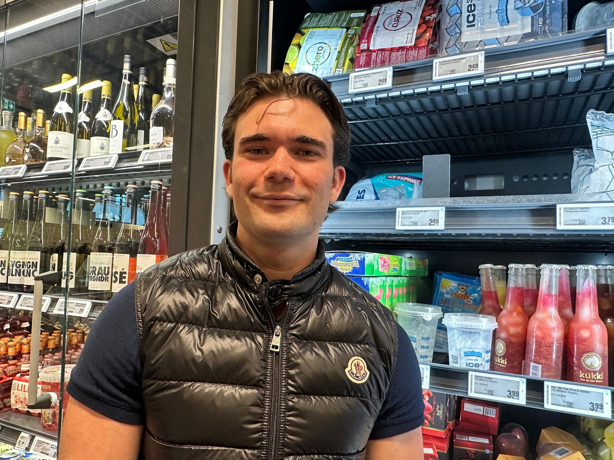 Ein junger Mann mit Steppweste und dunklem Haar steht vor einem Kühlregal. Hinter ihm sind Eiswürfel im Becher zu sehen, und allerlei andere Getränke.