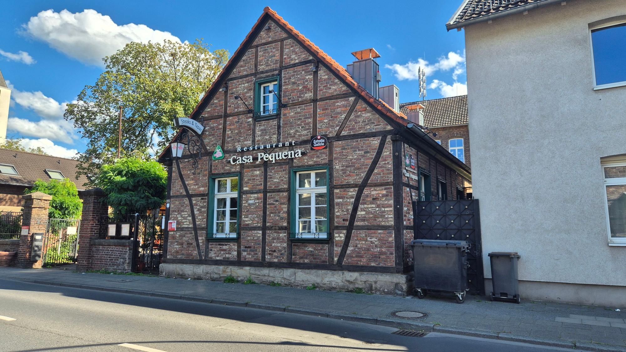 An einer Straße steht ein alter Ziegelbau mit Fachwerk-Elementen. Es gibt ein Tor, das zu einem Hof führt.