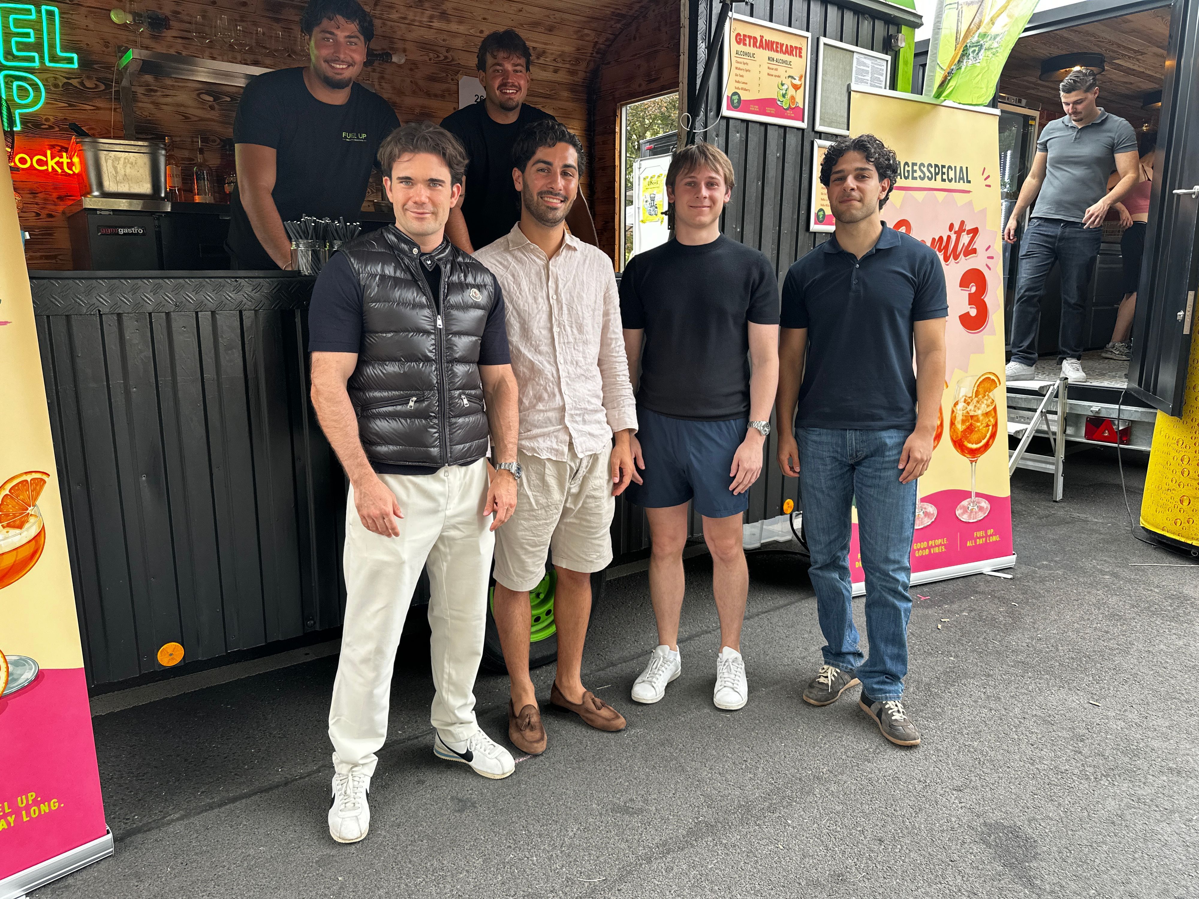 Vier junge Männer stehen vor einem mobilen Getränkestand, der Aperol Spritz verkauft.