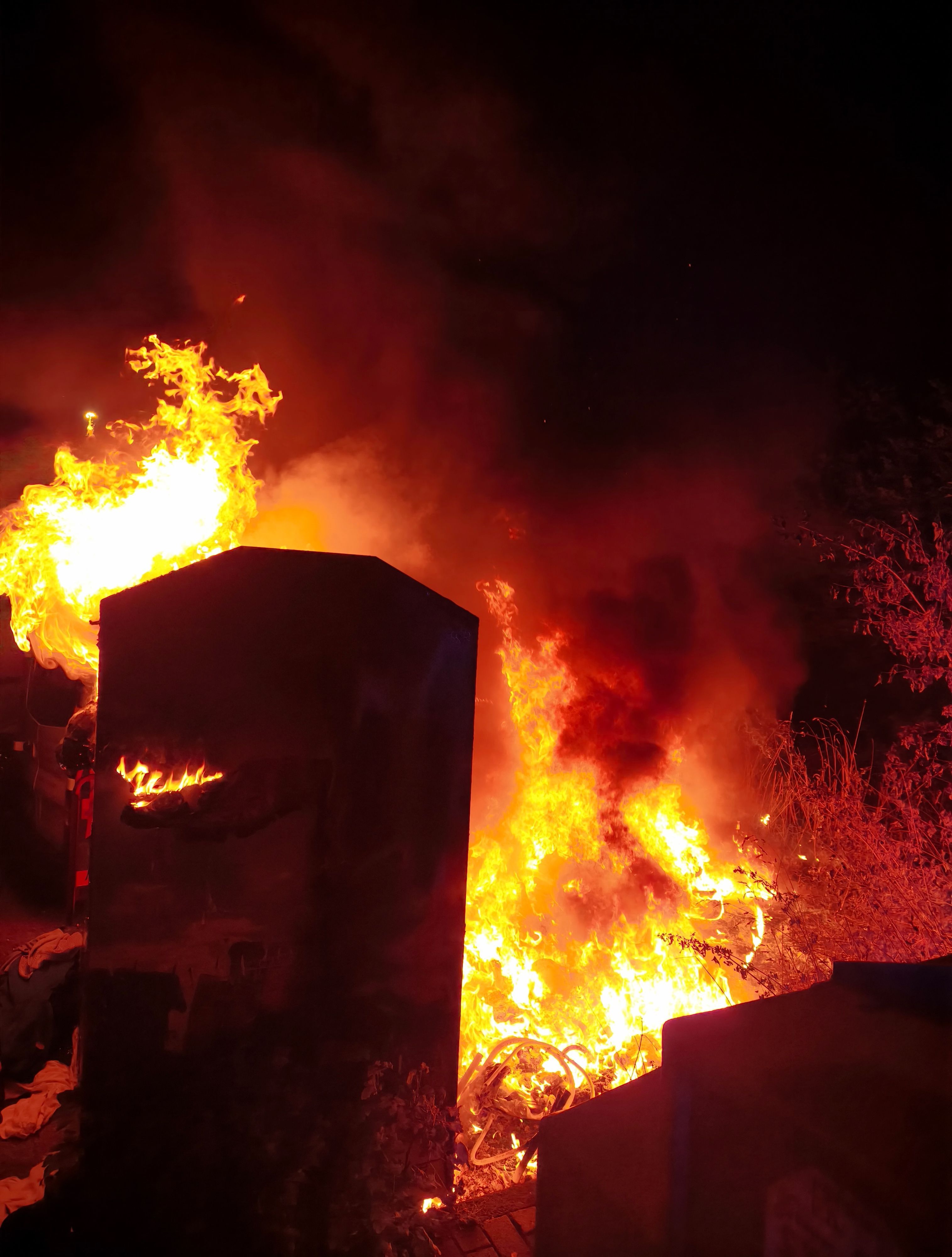 Ein Altkleidercontainer und die Grünfläche daneben stehen in Flammen.