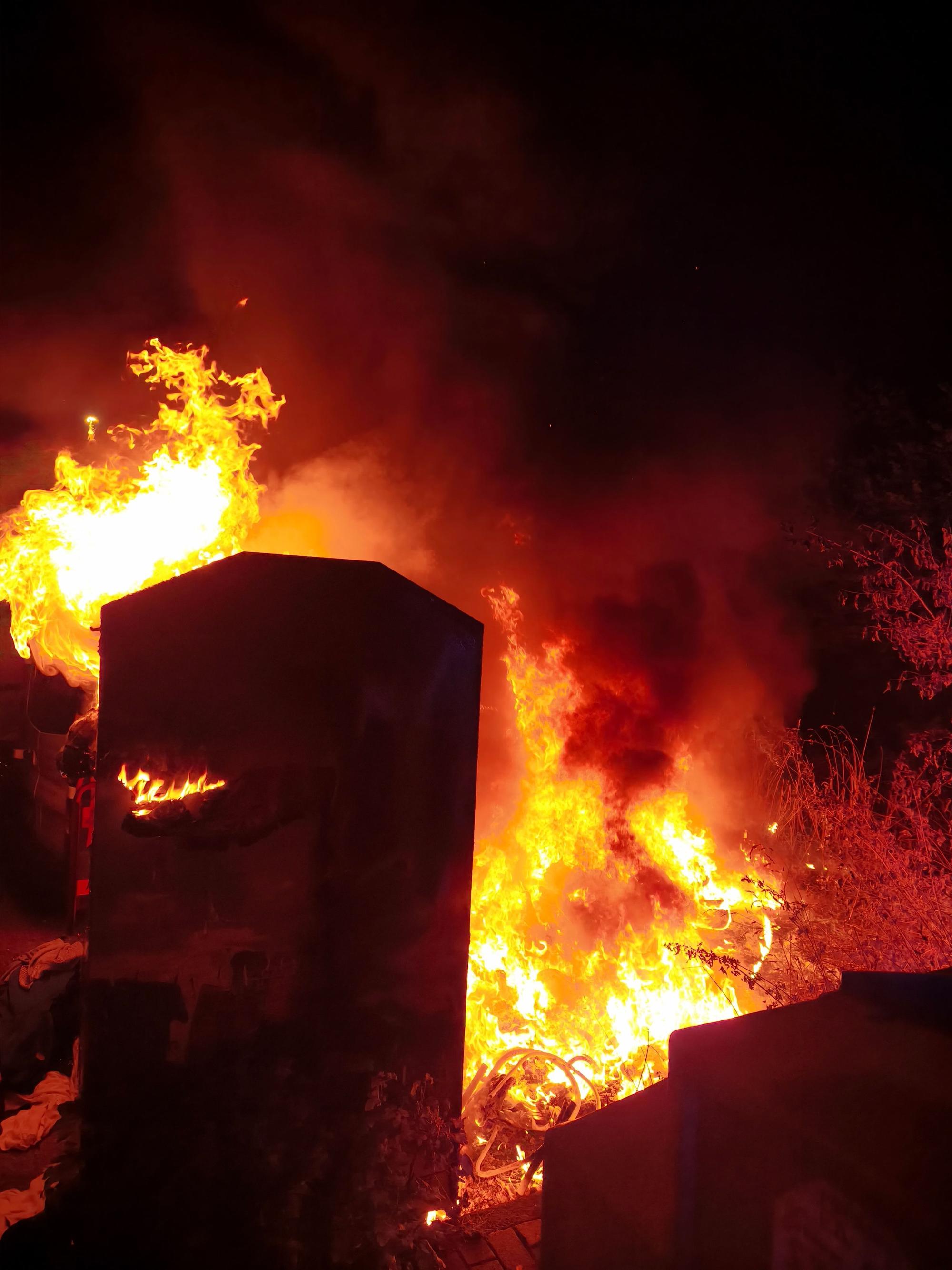 Ein Altkleidercontainer und die Grünfläche daneben stehen in Flammen.