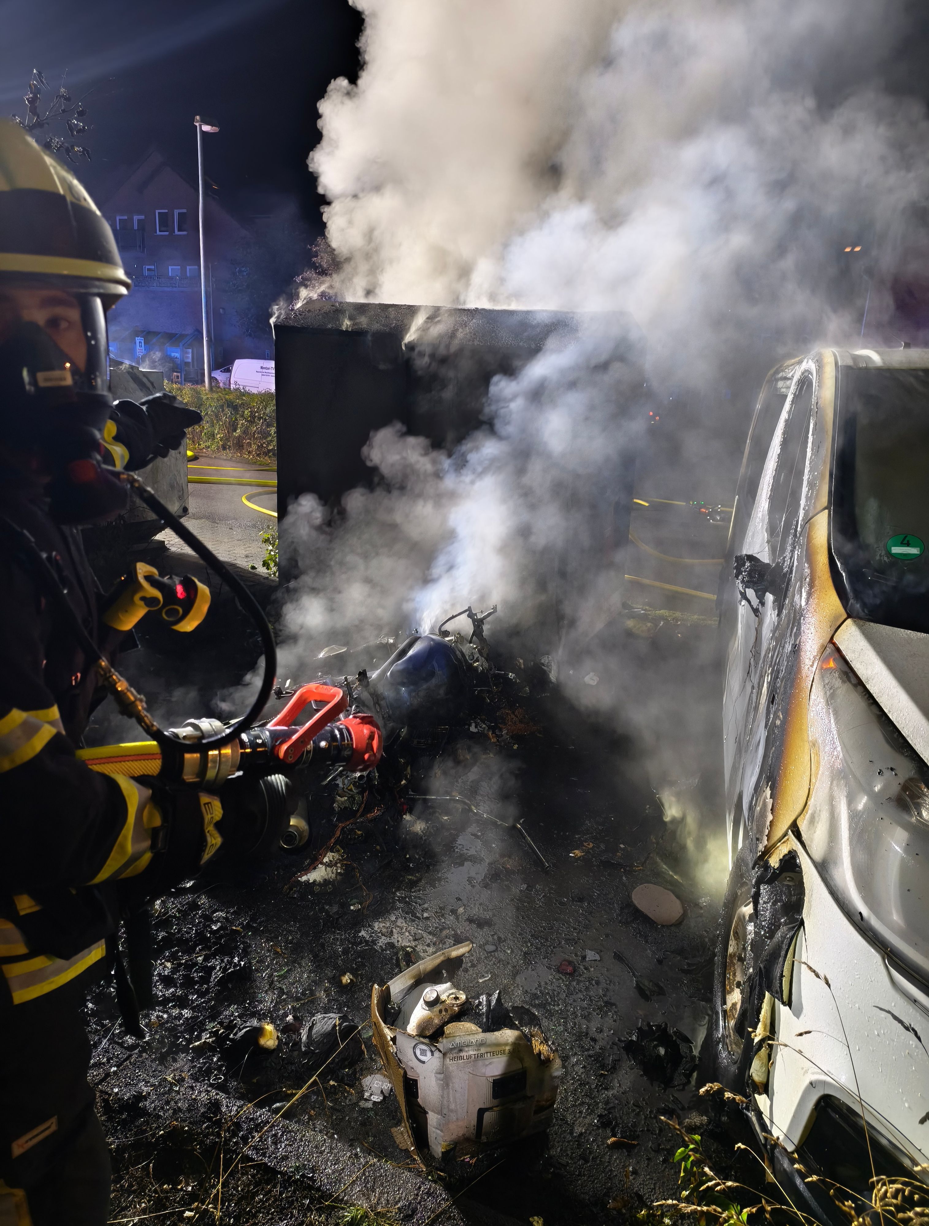 Ein Feuerwehrmann mit einem Strahlrohr steht neben einem verbrannten Altkleidercontainer und einem angeflämmten Auto.