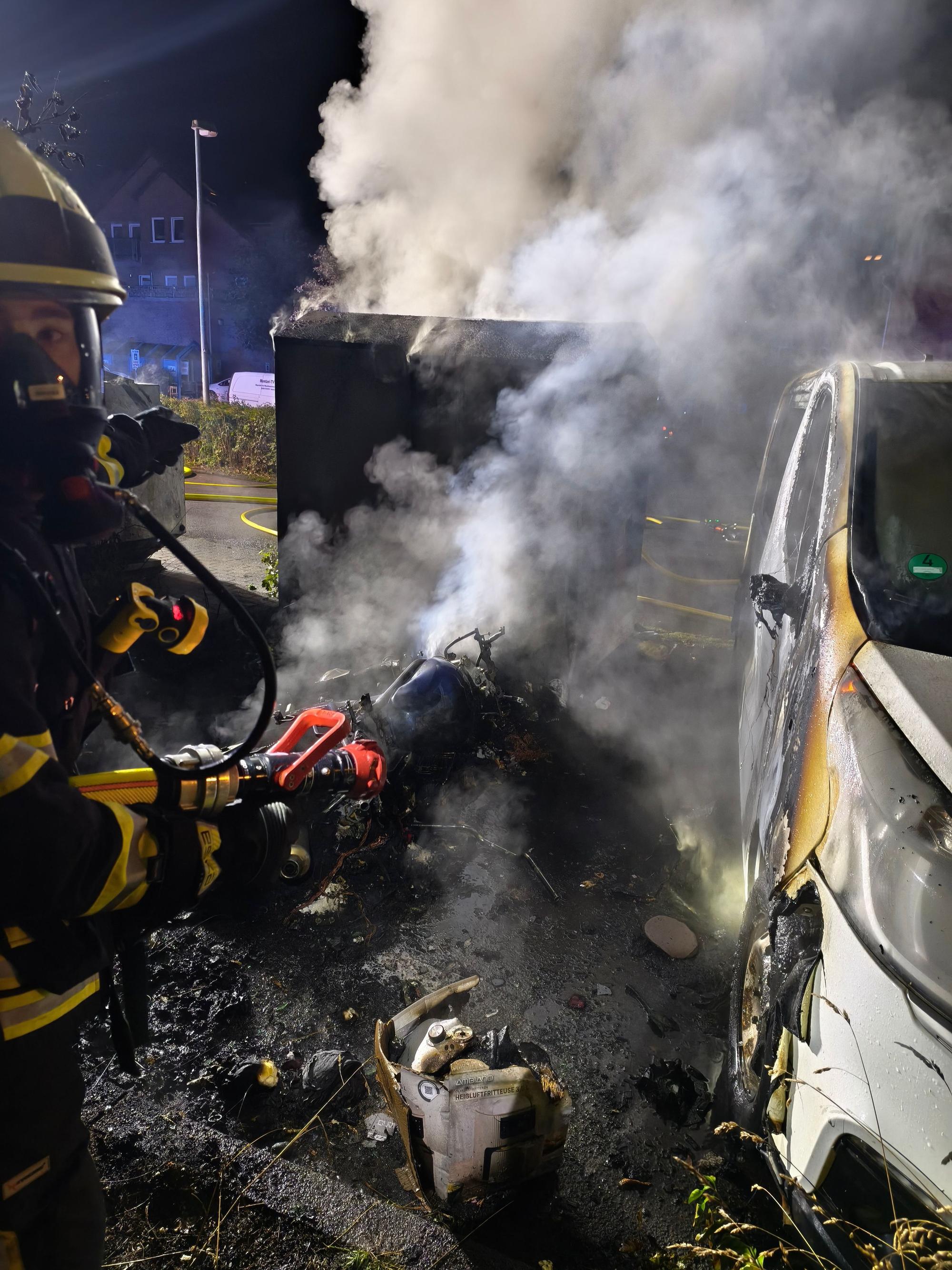 Ein Feuerwehrmann mit einem Strahlrohr steht neben einem verbrannten Altkleidercontainer und einem angeflämmten Auto.