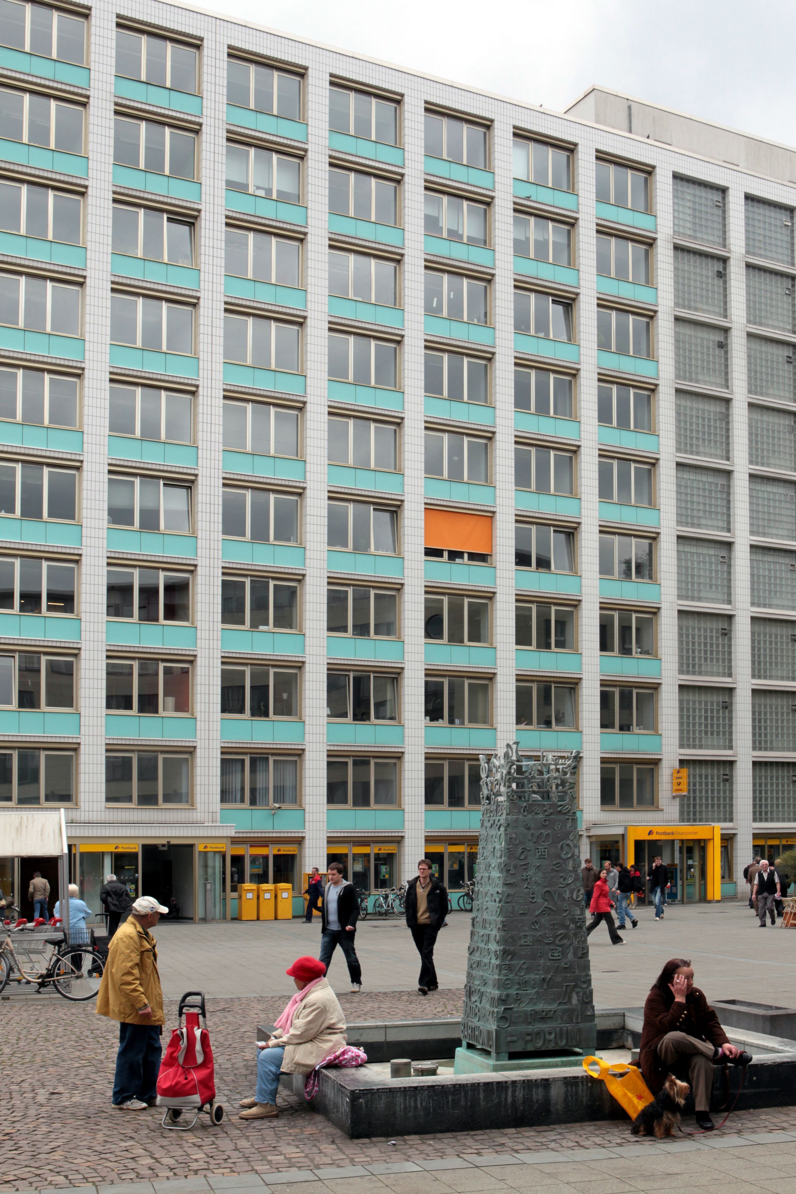 Auf einem Platz steht ein Brunnen, auf der Umrandung sitzen Menschen.