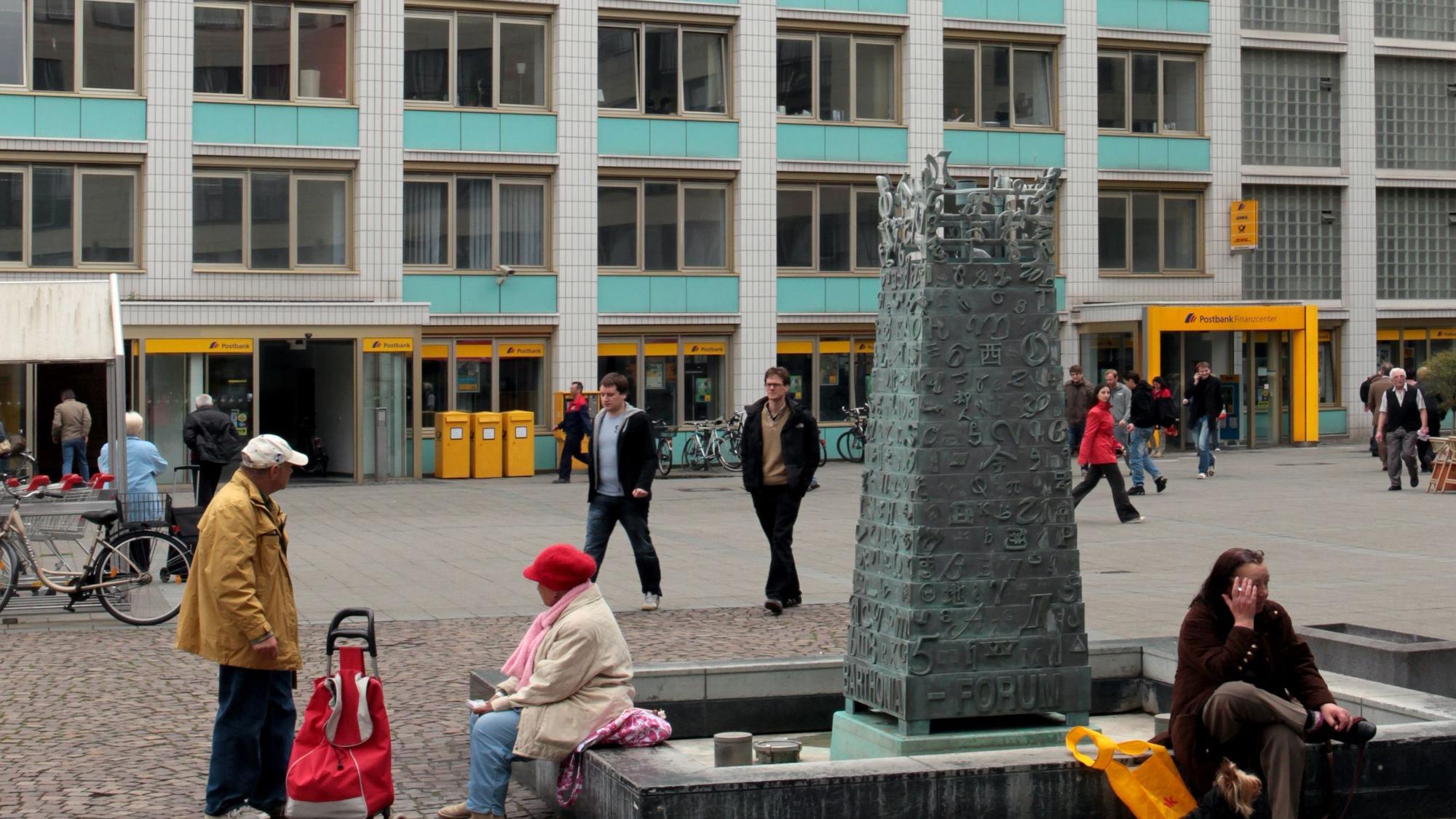 Auf einem Platz steht ein Brunnen, auf der Umrandung sitzen Menschen.