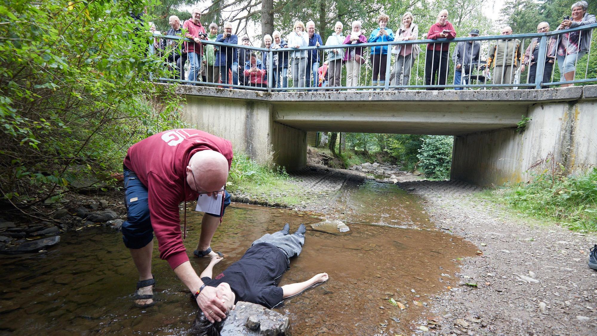 Ein Jugendlicher liegt in einem Bach, der sehr wenig Wasser führt, und wird vom Pfarrer getauft.