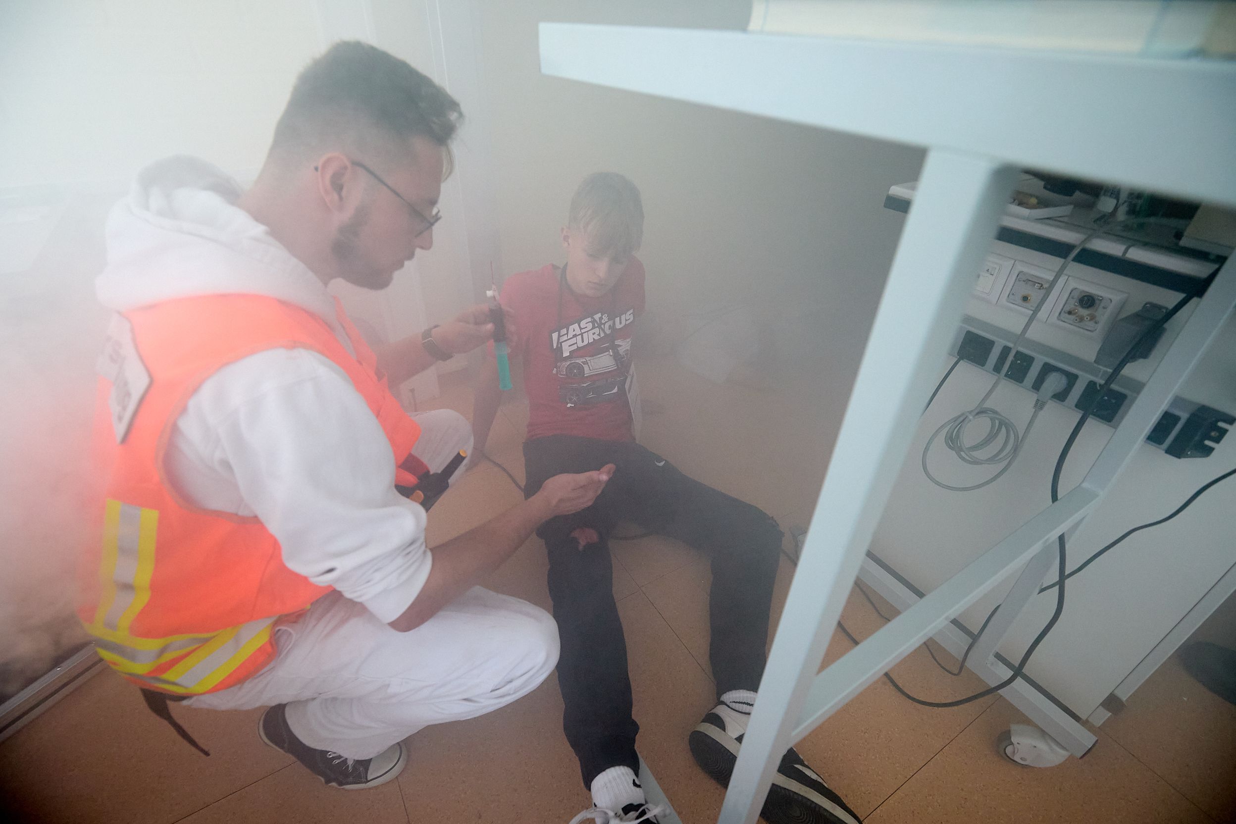 Ein Mann in orangefarbener Warnweste hockt in einem vernebelten Raum neben einem Jugendlichen.
