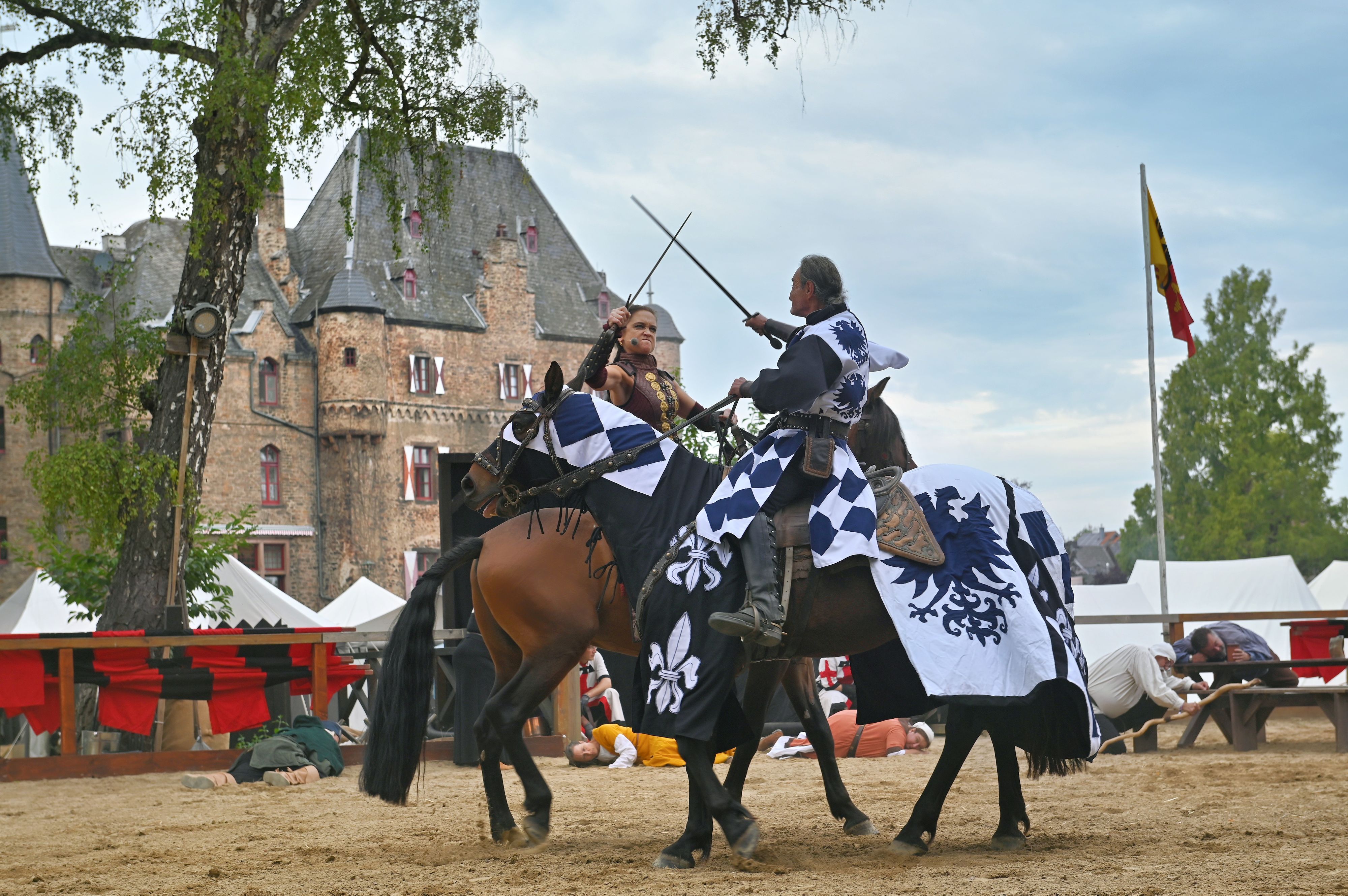 Eine Kriegerin und ein Ritter kämpfen mit Schwertern auf Pferden vor der Kulisse der Burg Satzvey.