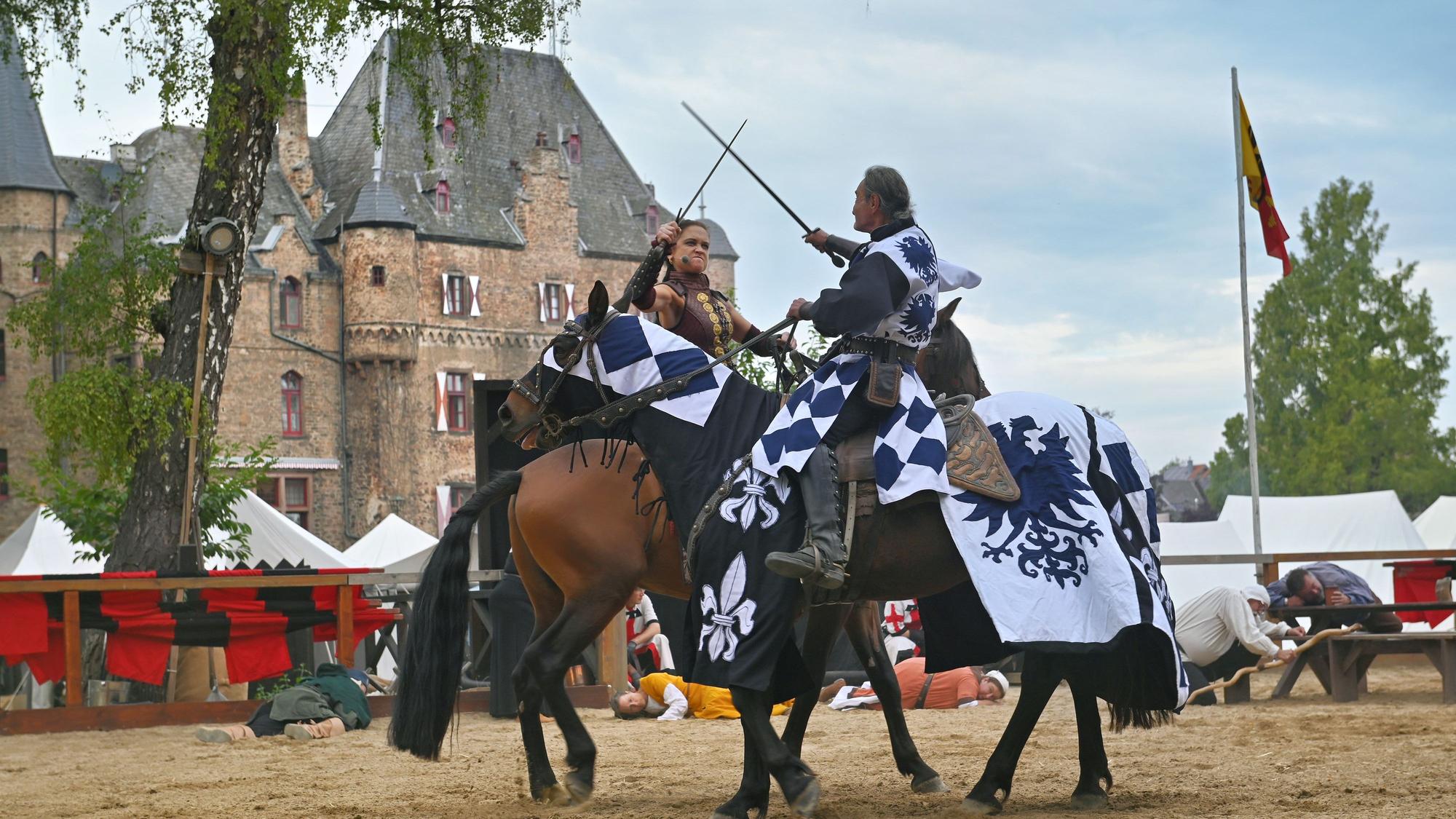 Eine Kriegerin und ein Ritter kämpfen mit Schwertern auf Pferden vor der Kulisse der Burg Satzvey.