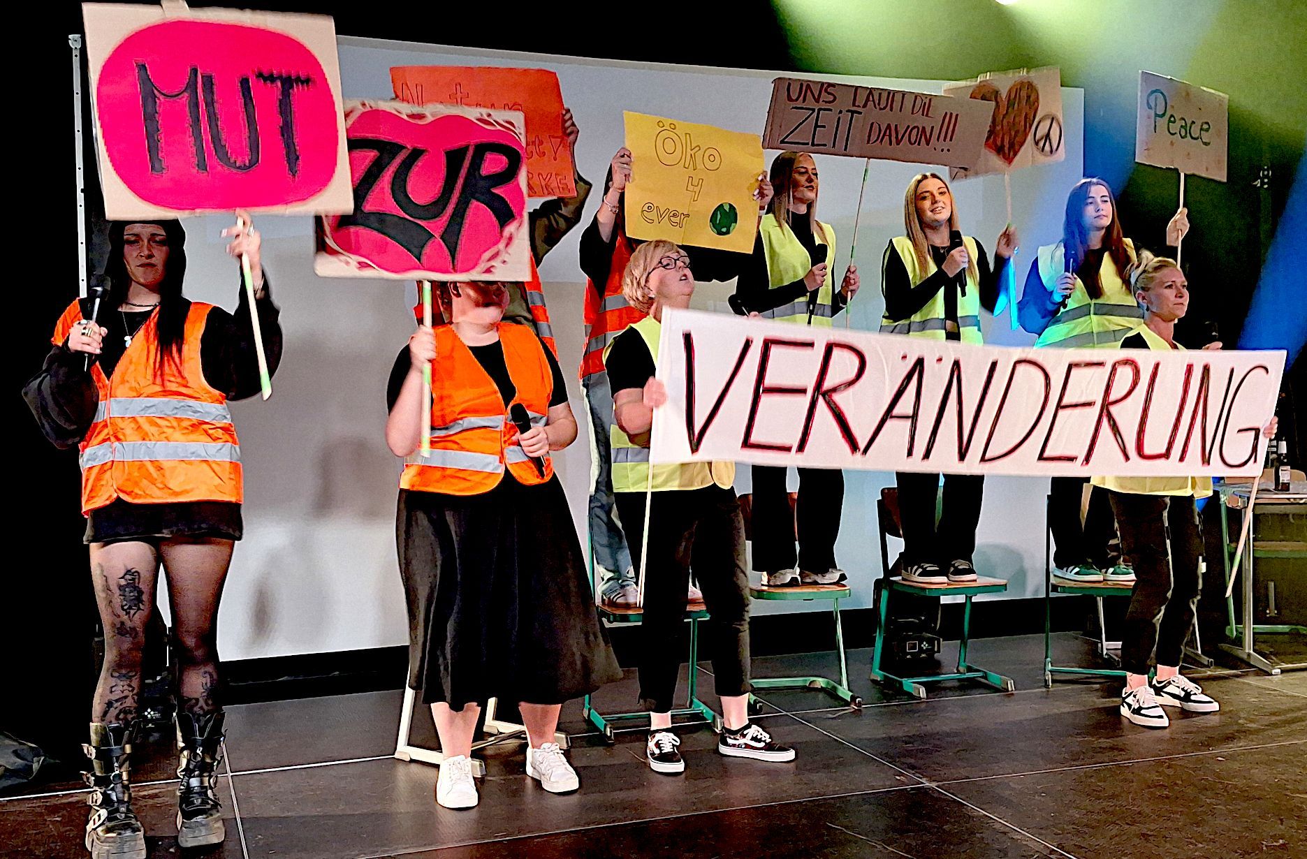 Mehre junge Frauen stehen in Warnwesten auf einer Bühne und halten Schilder in der Hand.
