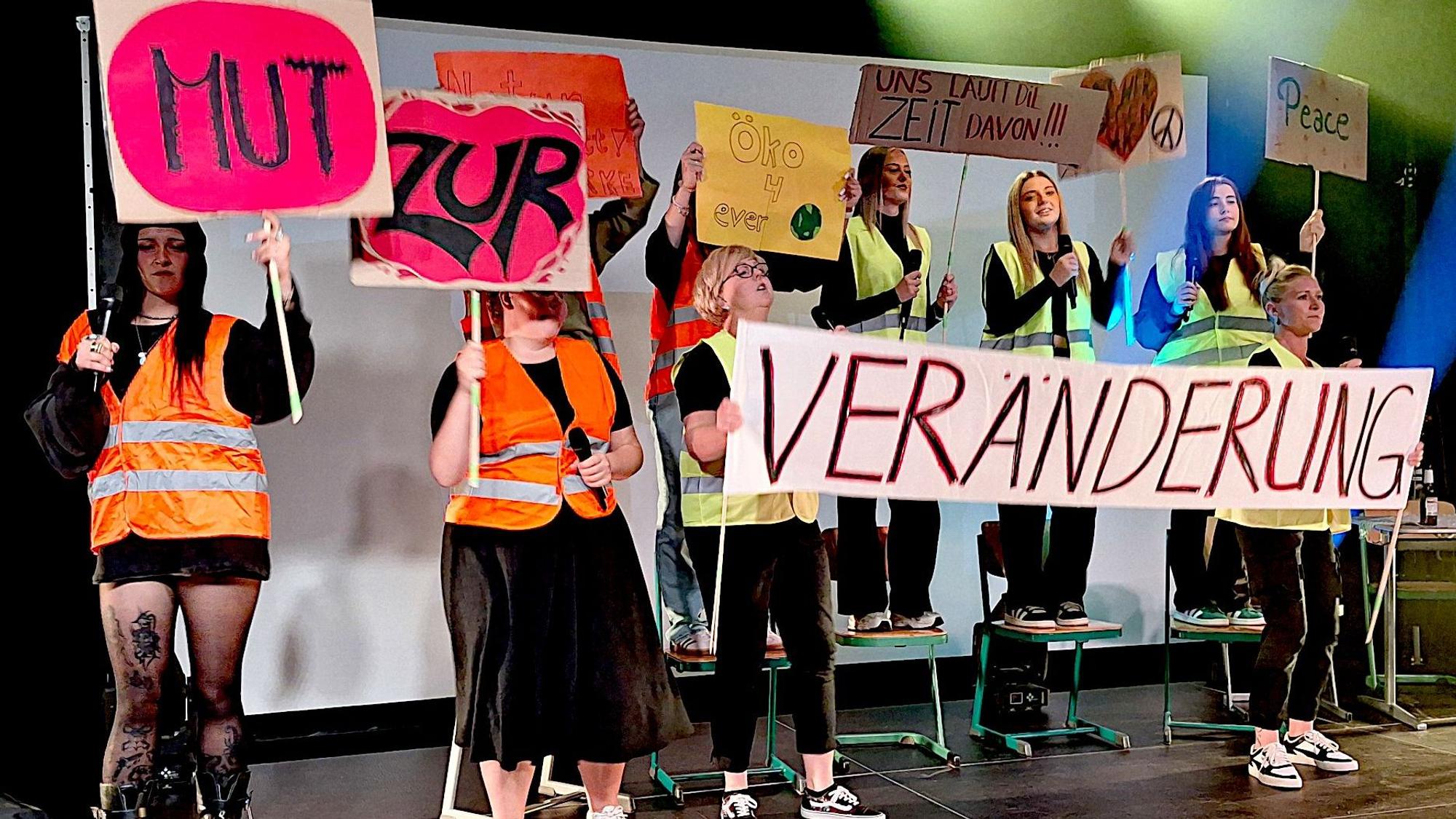 Mehre junge Frauen stehen in Warnwesten auf einer Bühne und halten Schilder in der Hand.