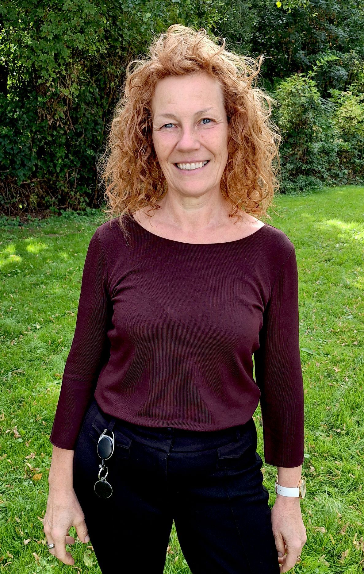 Die Frau in einem dunkelroten Oberteil und roten Locken steht auf einer Wiese.