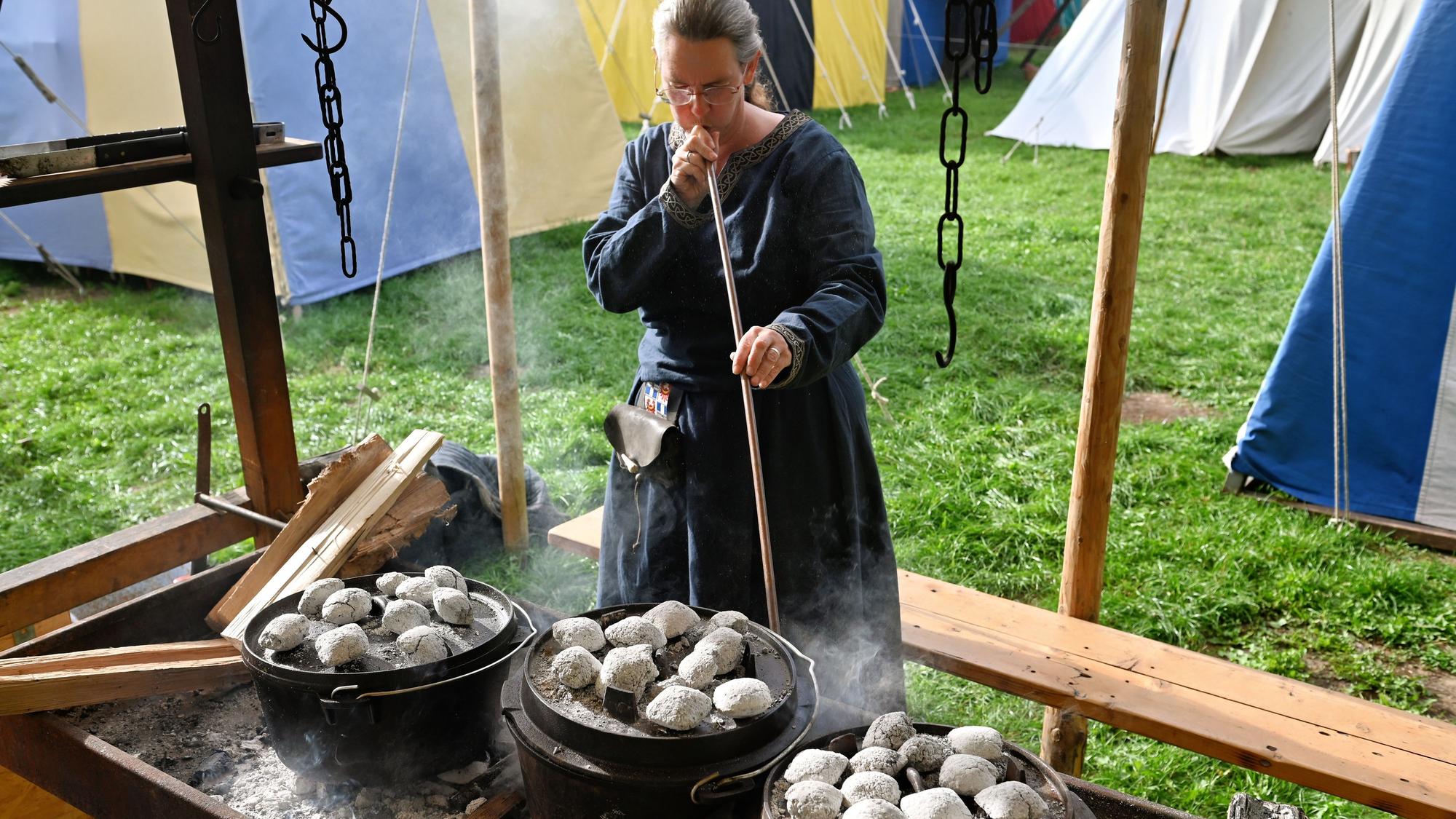Eine Frau in mittelalterlichem Gewand steht vor einem Gestell, auf dem drei gusseiserne Kochtöpfe stehen.