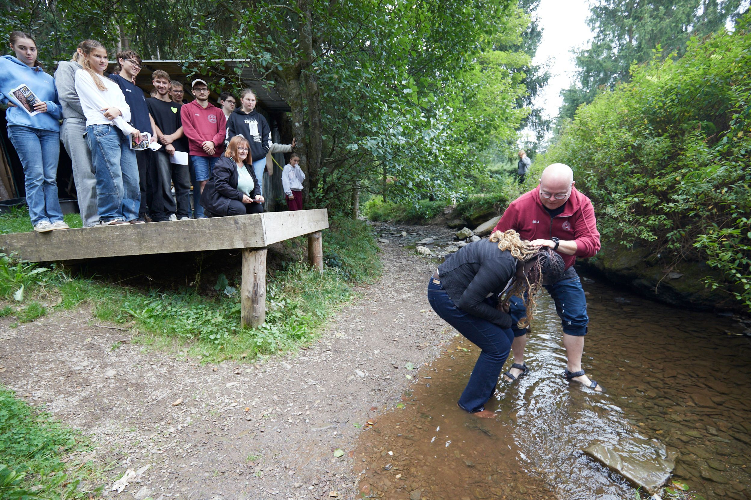 Die junge Frau steht in einem Bach und wird vom Pfarrer getauft, derr Wasser über ihren Kopf schüttet.