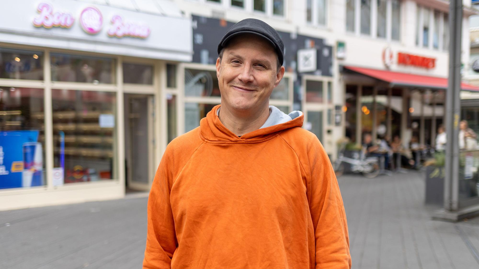 Ein Mann im orangefarbenen Pullover lächelt in die Kamera.