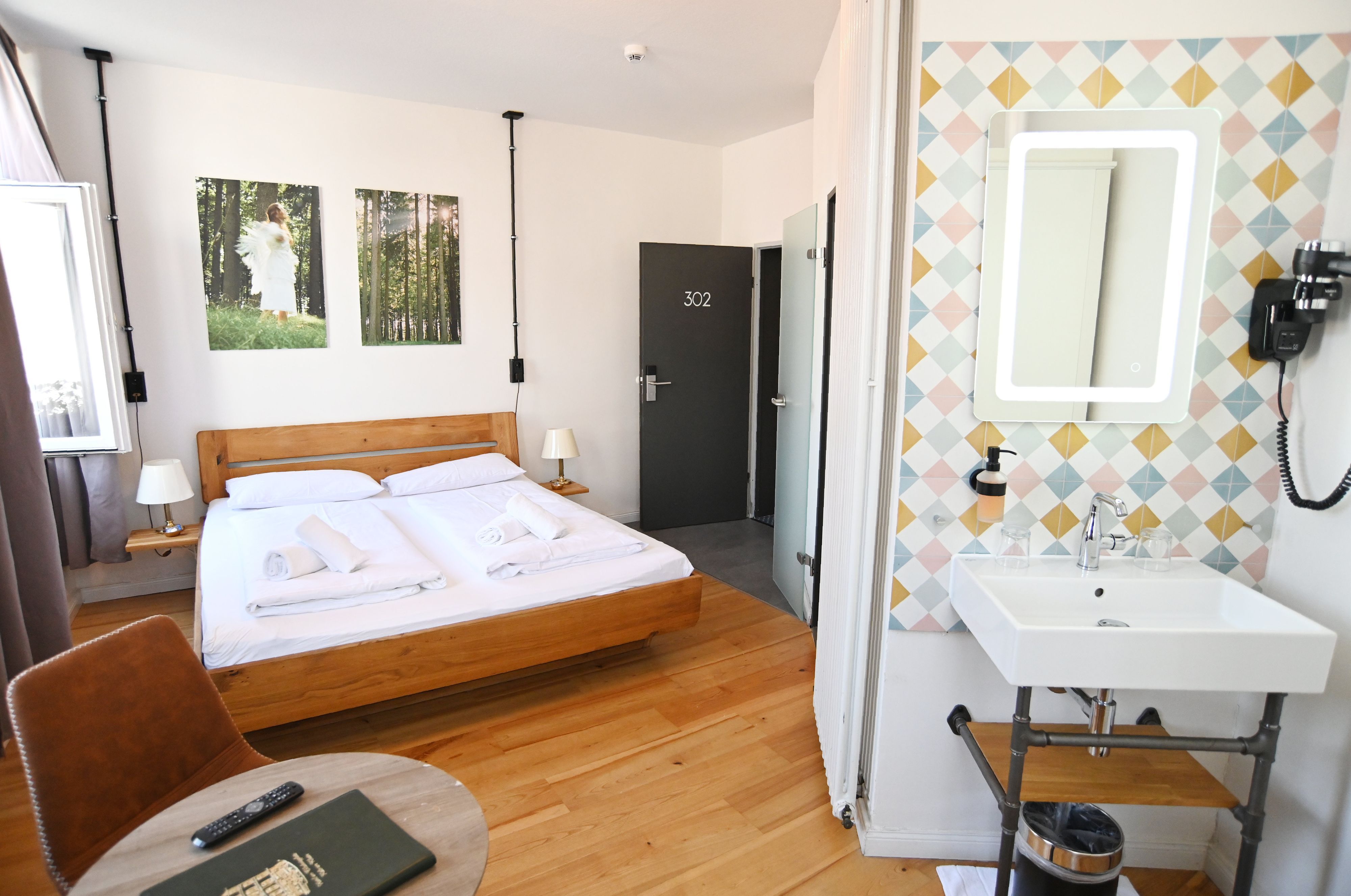 In einem hellen Zimmer steht ein Bett, in einer Nische ist ein Waschbecken, darunter ein Toiletten-Regal.